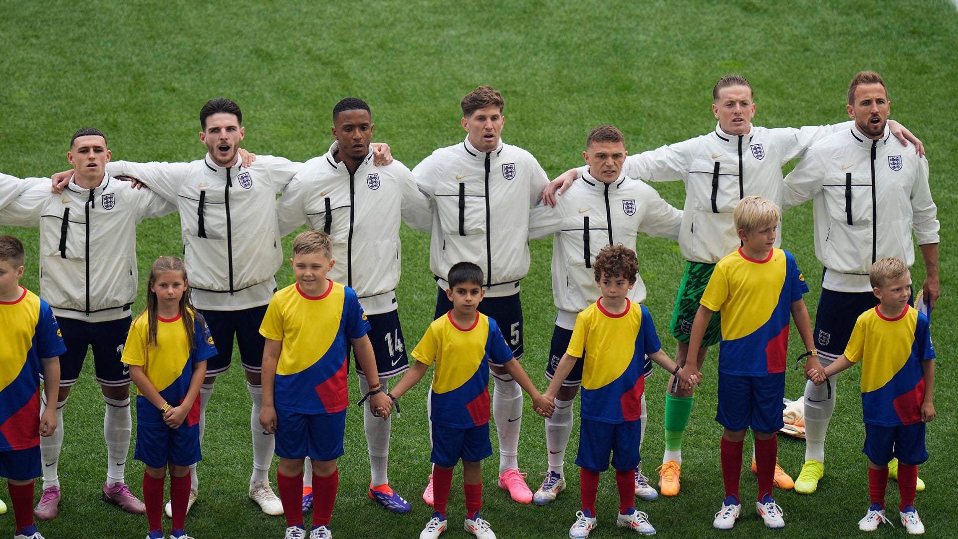Fotbollslag uppställt på gräsplan. Vuxna spelare i vita jackor och unga ungdomar i gula och blå fotbollströjor står tillsammans i en rad.