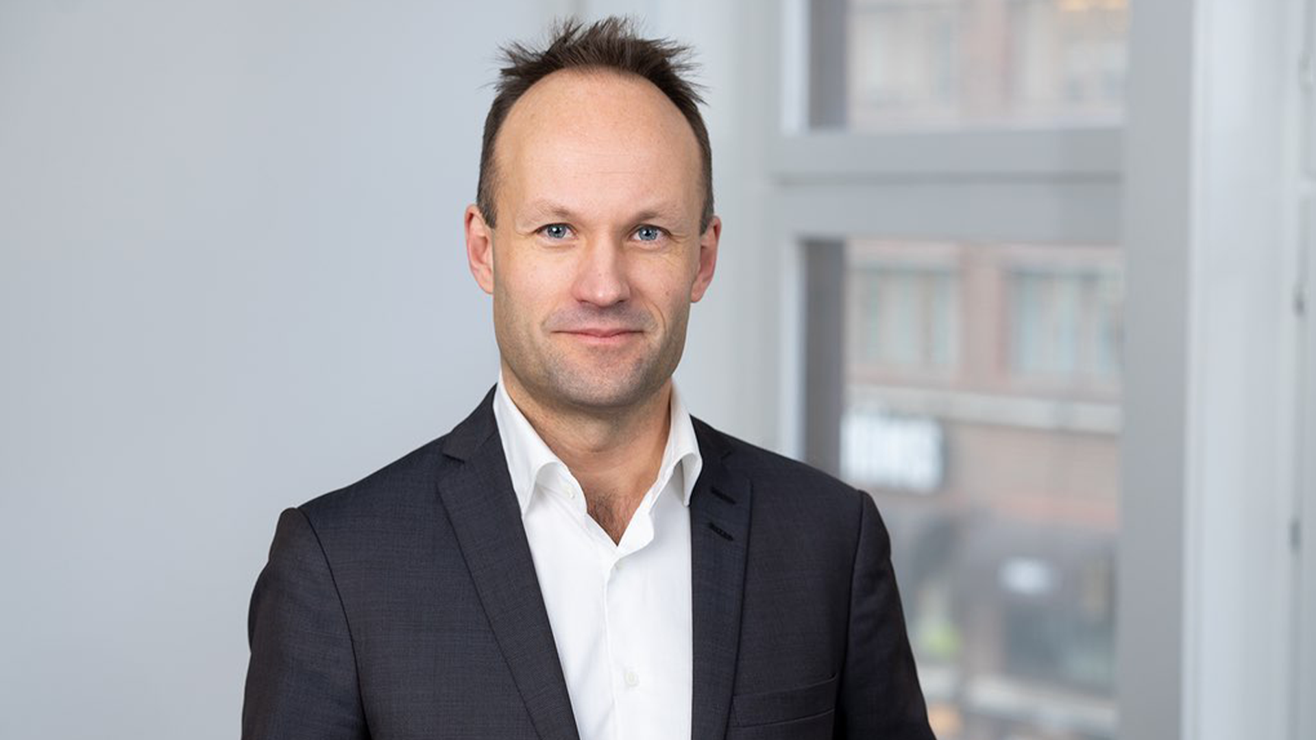 Porträtt av en man i mörk kostym och vit skjorta framför ett fönster.