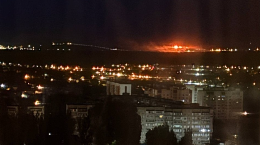 Nattfotografi av en stad med huslampor och gatlyktor. En stor brand eller explosion syns i bakgrunden med orange glöd på himlen.
