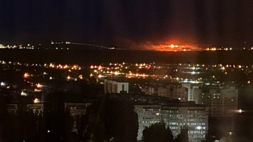 Nattfotografi av en stad med huslampor och gatlyktor. En stor brand eller explosion syns i bakgrunden med orange glöd på himlen.