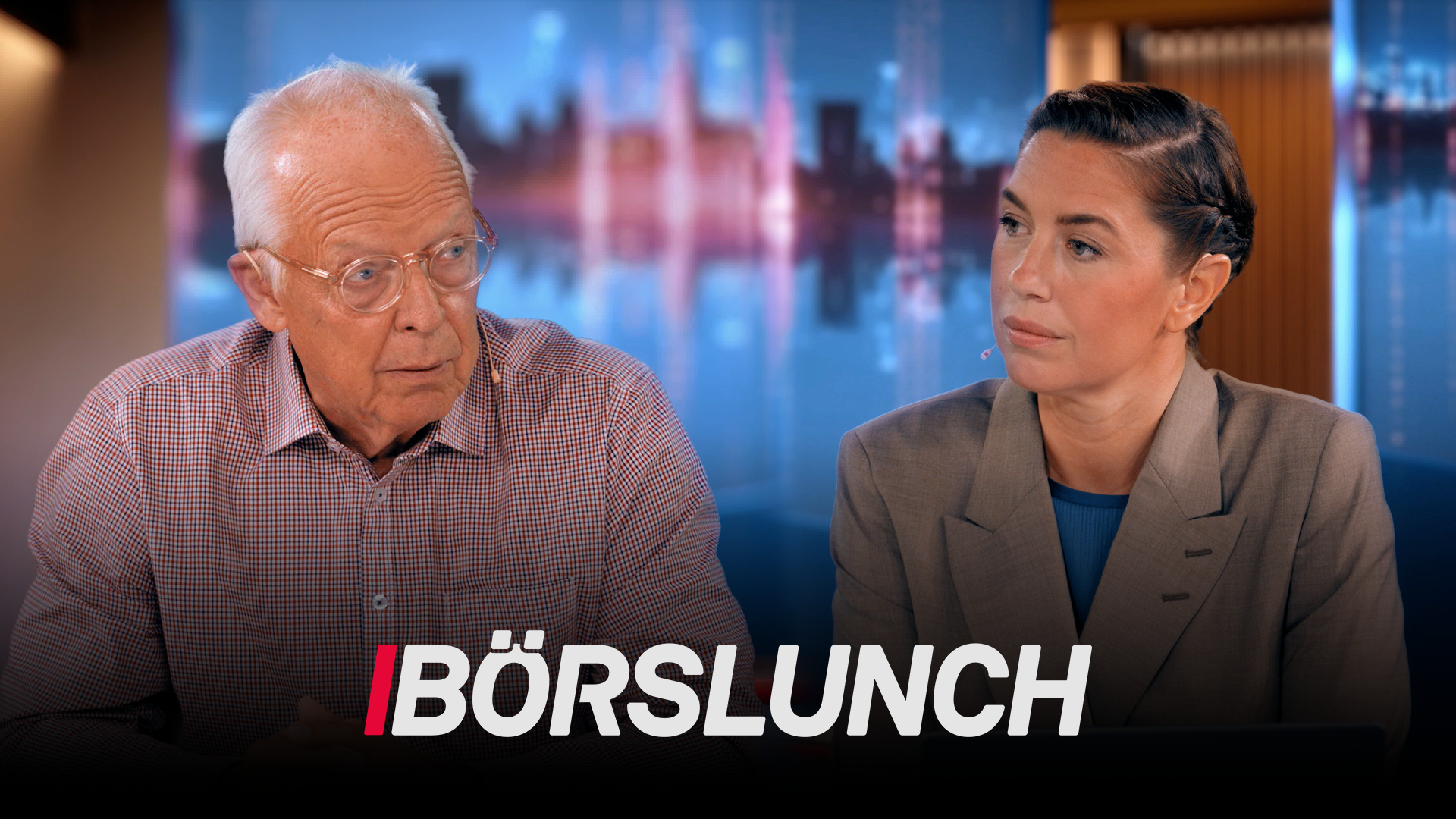 En äldre man och en kvinna sitter vid ett studiobord under ett TV-program kallat Börslunch. I bakgrunden syns en stadshorisont med blå och röd belysning.