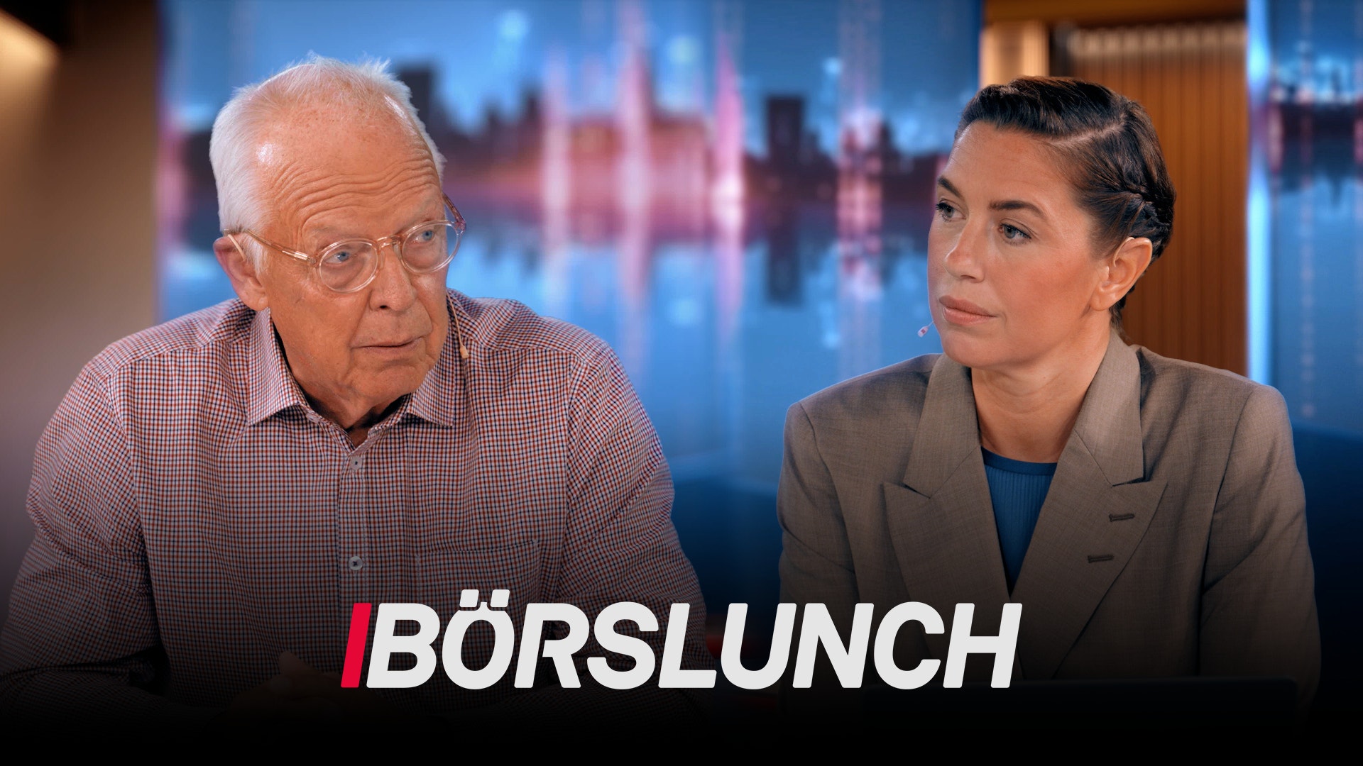 En äldre man och en kvinna sitter vid ett studiobord under ett TV-program kallat Börslunch. I bakgrunden syns en stadshorisont med blå och röd belysning.