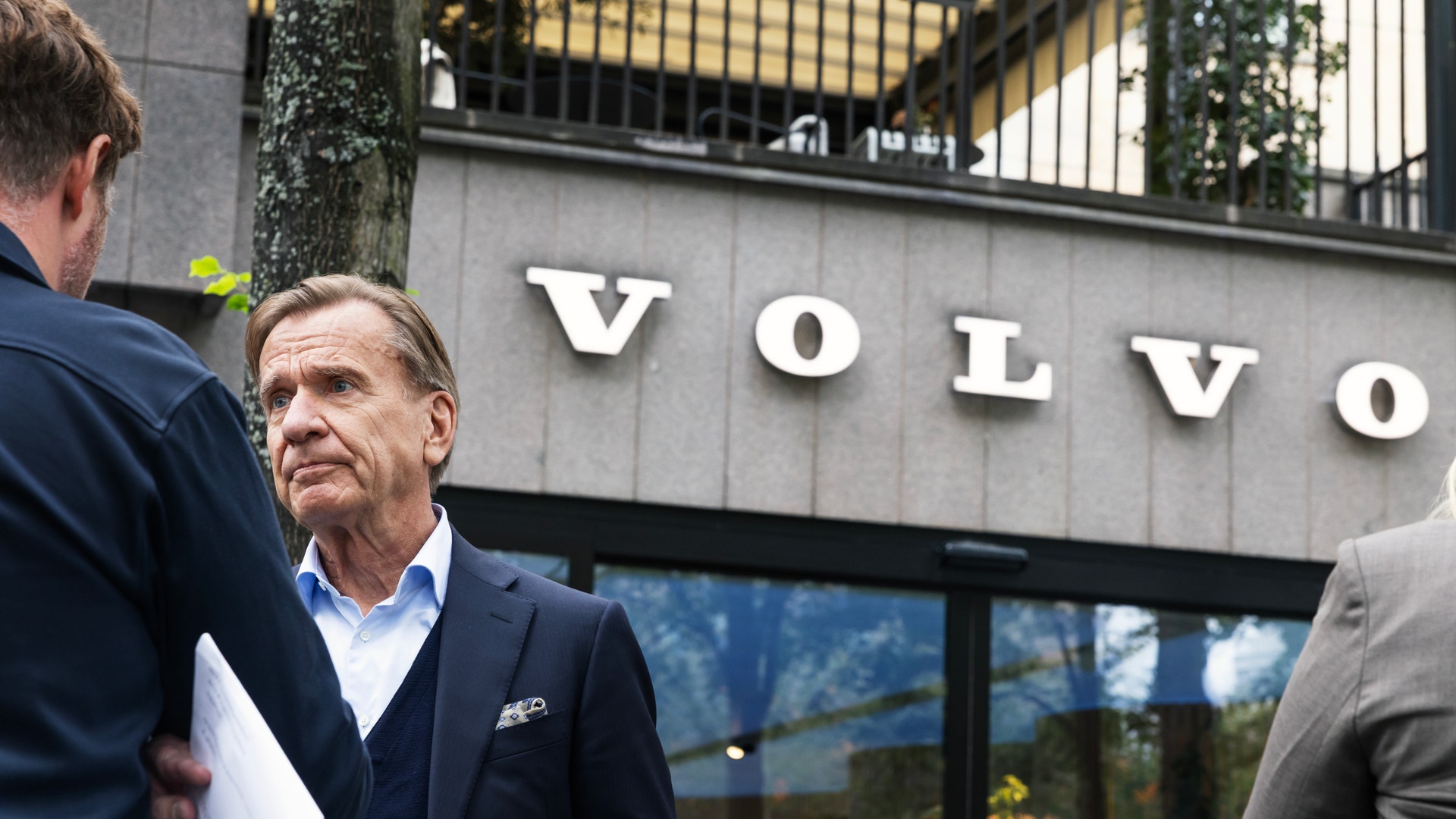 Håkan Samuelsson, vd för Volvo Cars, står utomhus framför en byggnad med Volvos logotyp och tittar allvarligt åt sidan.