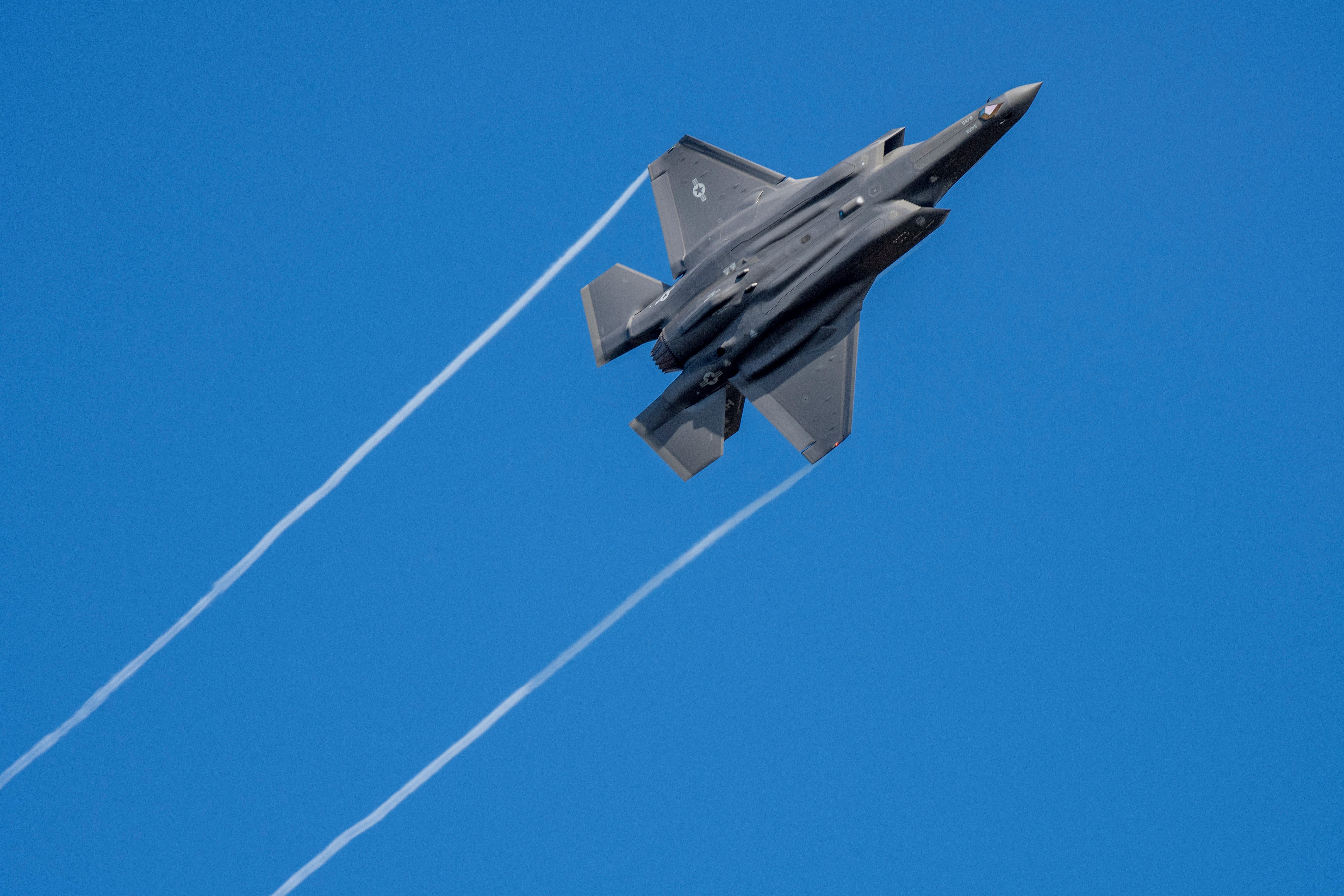 Ett grått stridsflygplan av typen F-35 sett underifrån när det stiger mot en klarblå himmel och lämnar två vita ränder efter sig.
