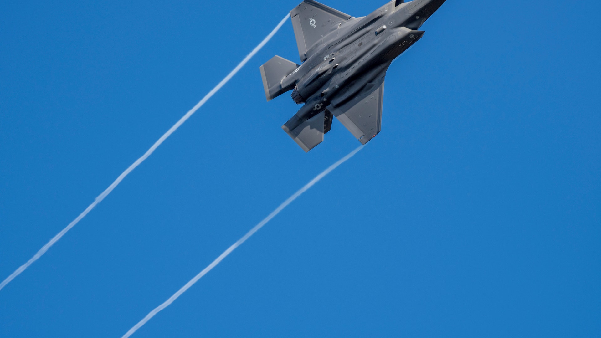 Ett grått stridsflygplan av typen F-35 sett underifrån när det stiger mot en klarblå himmel och lämnar två vita ränder efter sig.