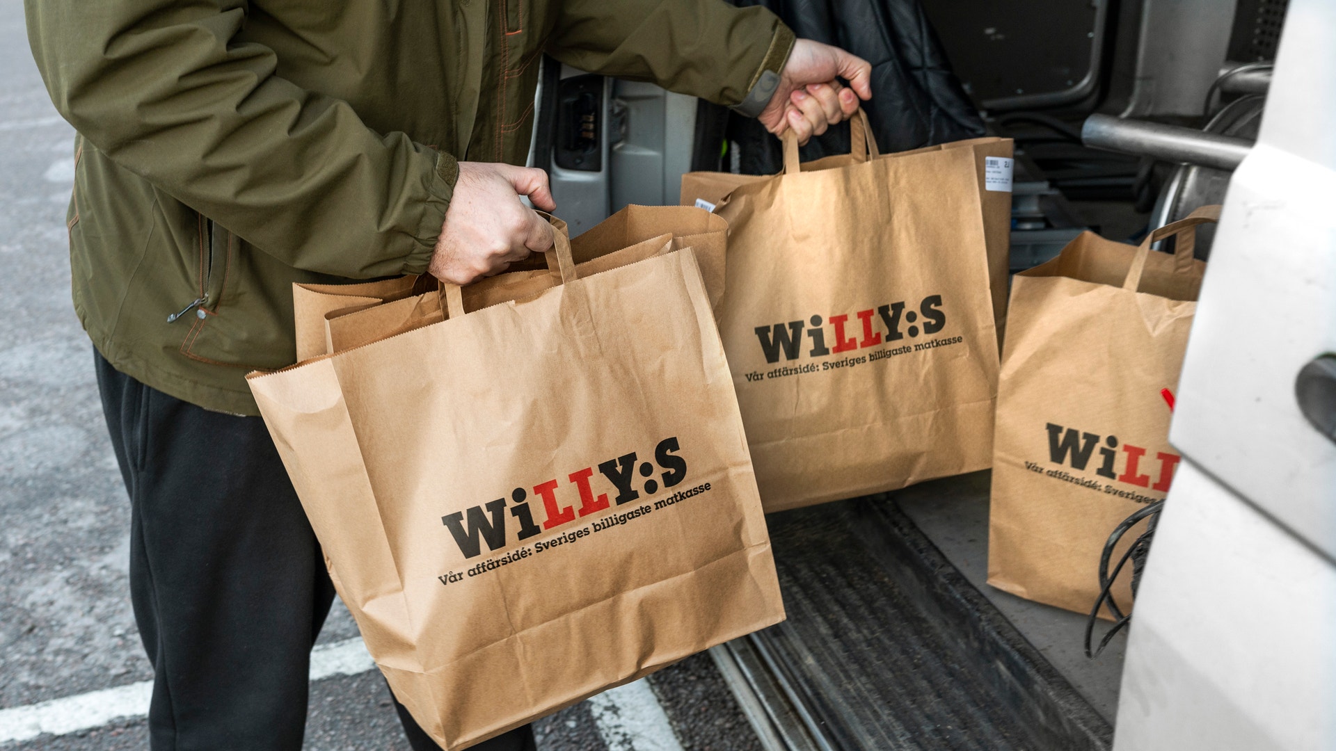 En person håller tre papperspåsar märkta WILLY:S framför en öppen bildörr.