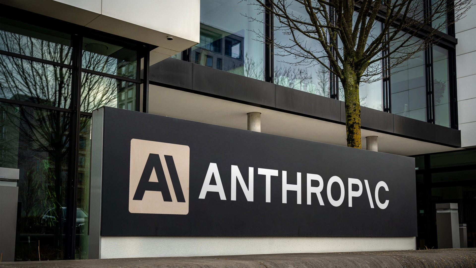 Skylt med Anthropic-logotypen på en modern byggnad med stora fönster och träd i bakgrunden.