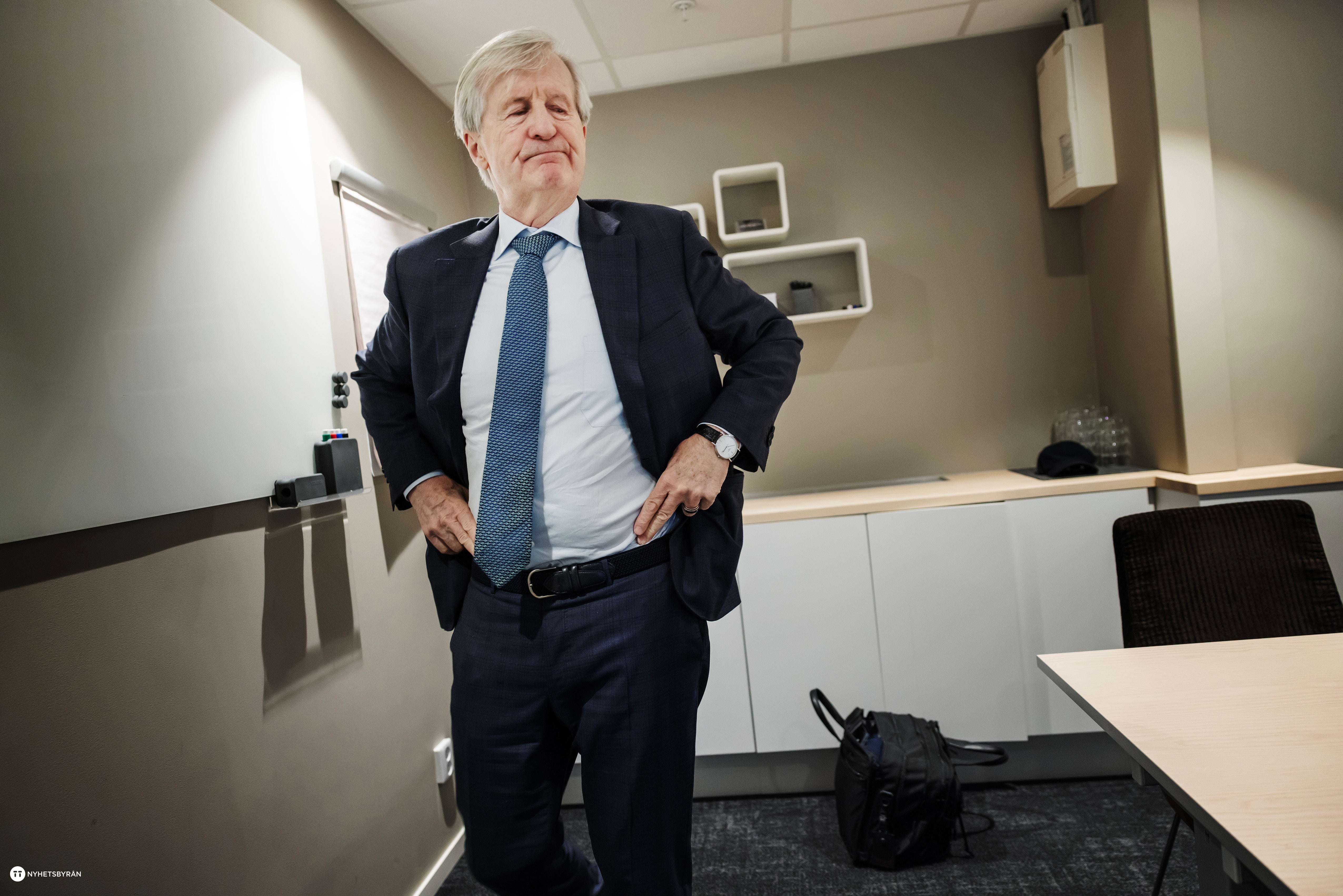 Äldre man i mörk kostym och blå slips står i ett modernt kontorsrum med händerna i byxfickorna.