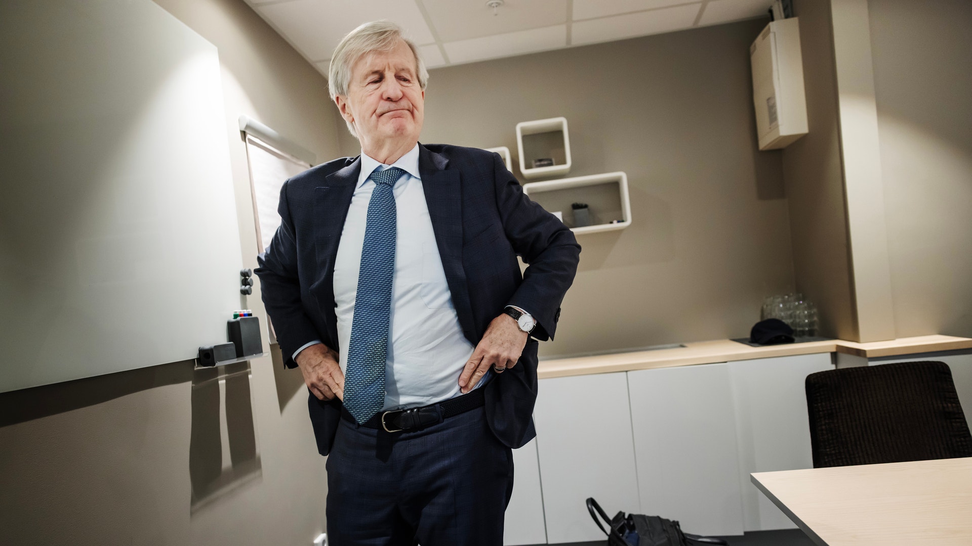 Äldre man i mörk kostym och blå slips står i ett modernt kontorsrum med händerna i byxfickorna.