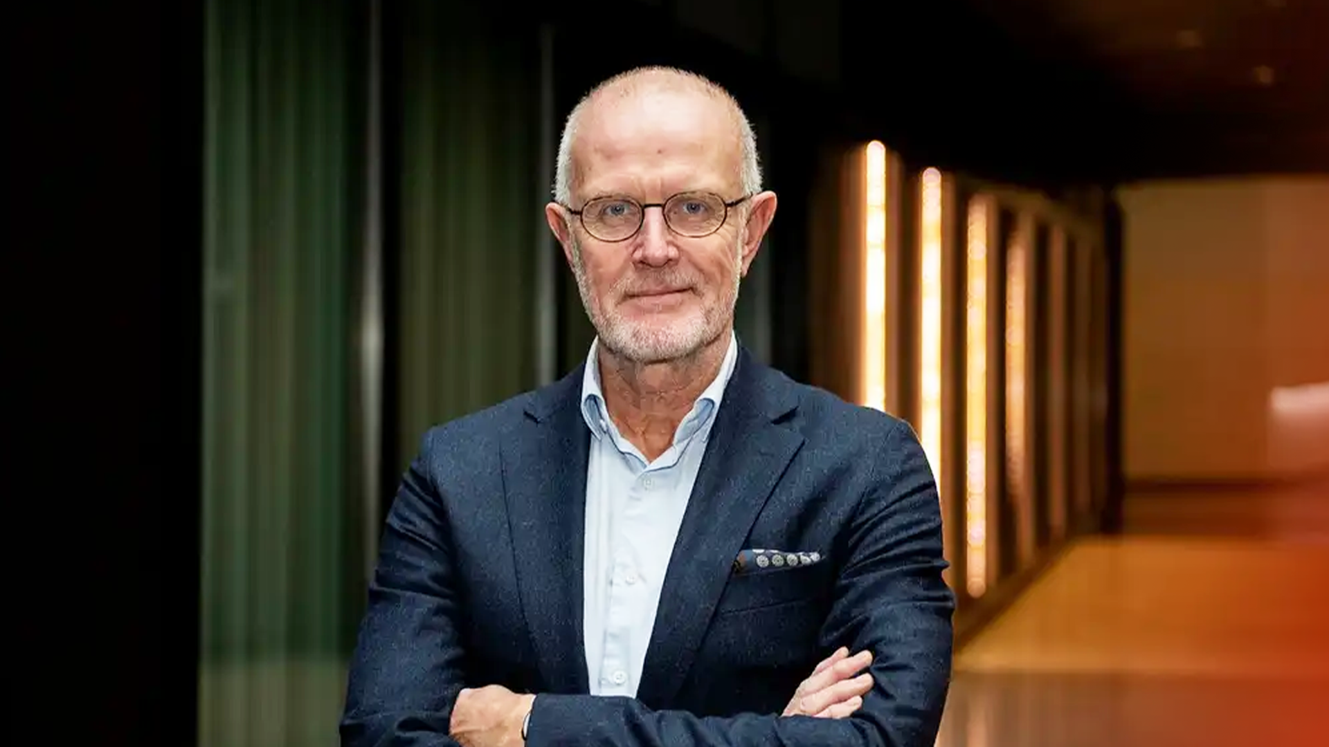 Porträttfoto av en äldre man med glasögon och kort grått hår, klädd i en mörkblå kavaj över en ljusblå skjorta. Han står med armarna i kors framför en modern bakgrund med gröna gardiner och varm belysning från gula vägglamper.