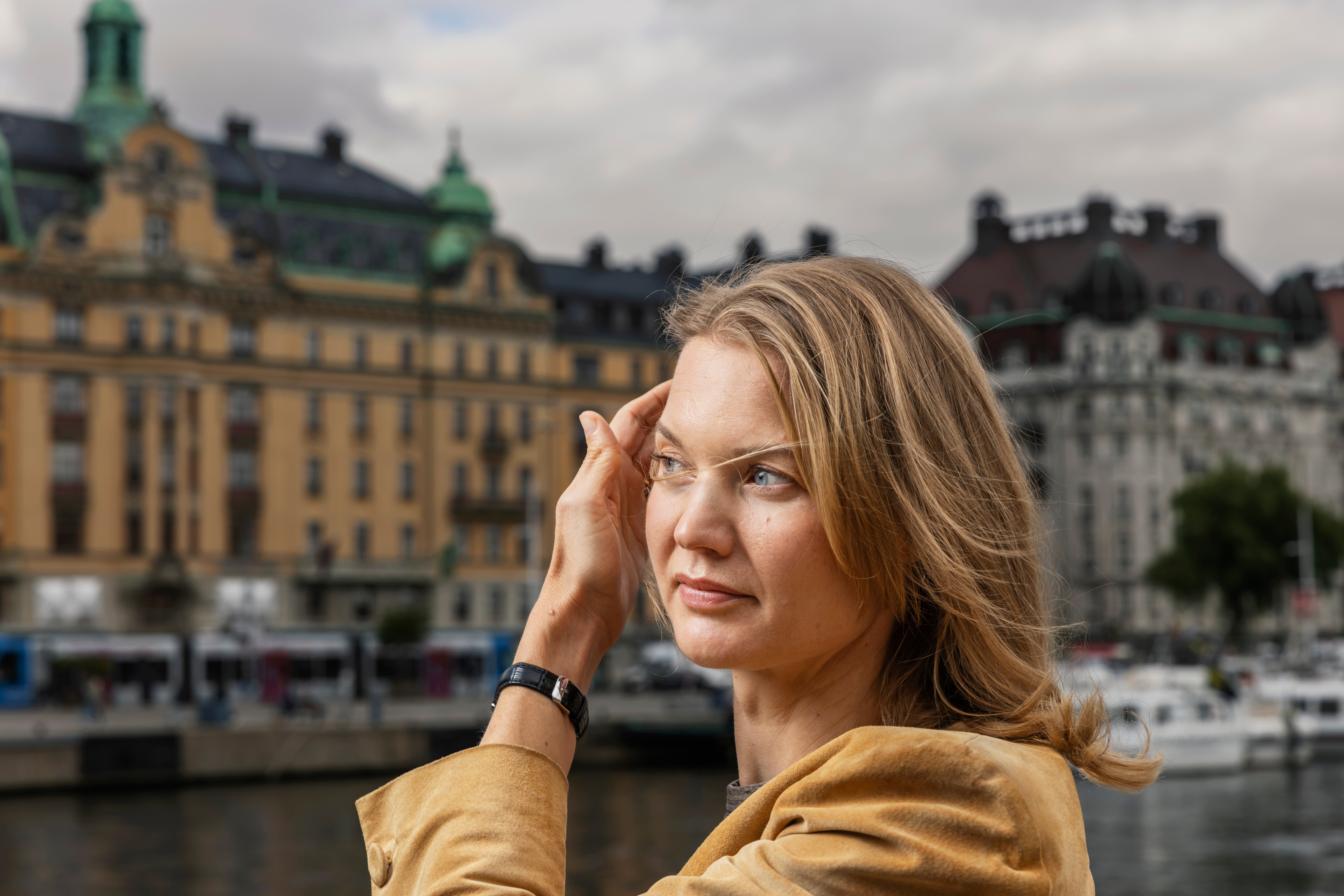 Porträtt av en kvinna i gul jacka som står framför klassiska byggnader vid en vattenkant.
