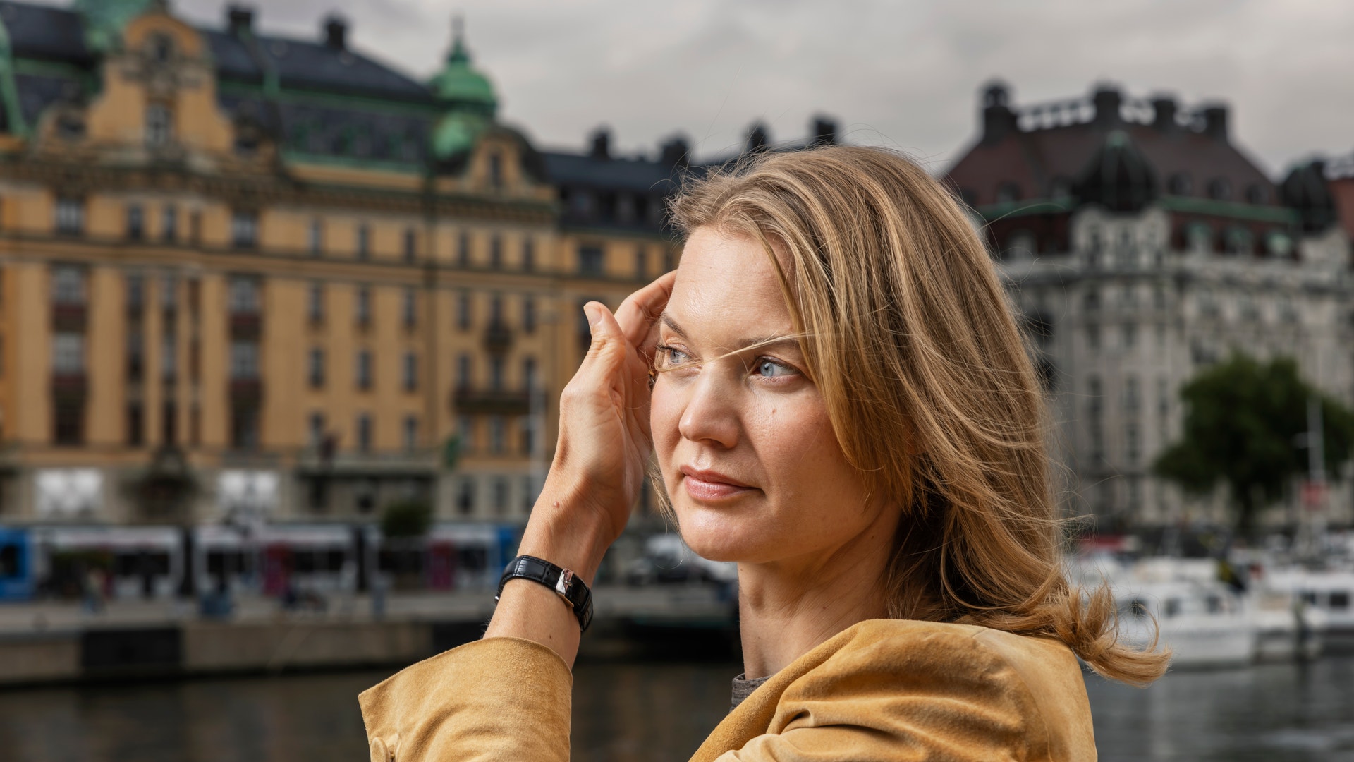 Porträtt av en kvinna i gul jacka som står framför klassiska byggnader vid en vattenkant.