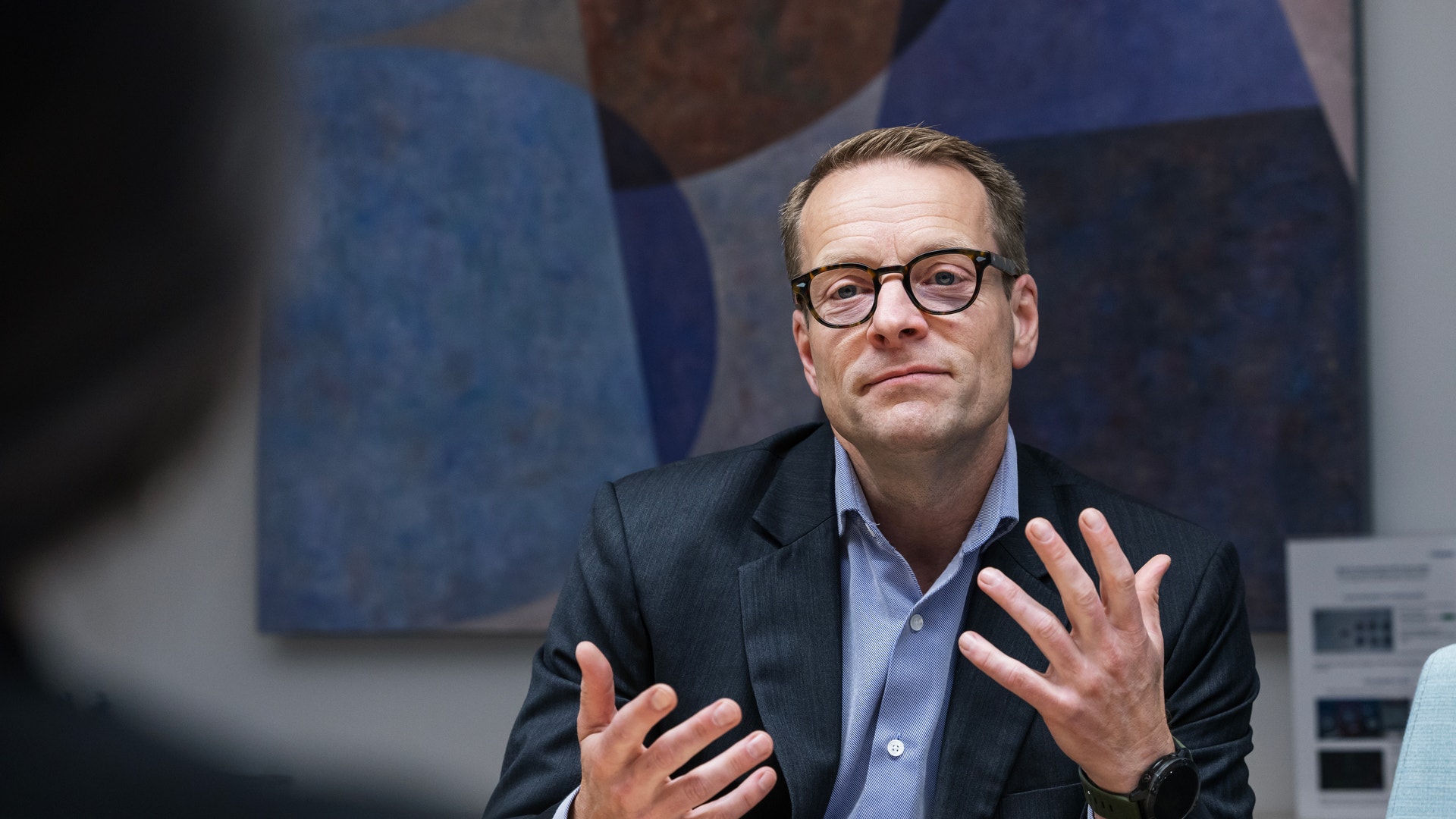 Man i glasögon och mörk blazer gestikulerar medan han talar i en kontorsmiljö.
