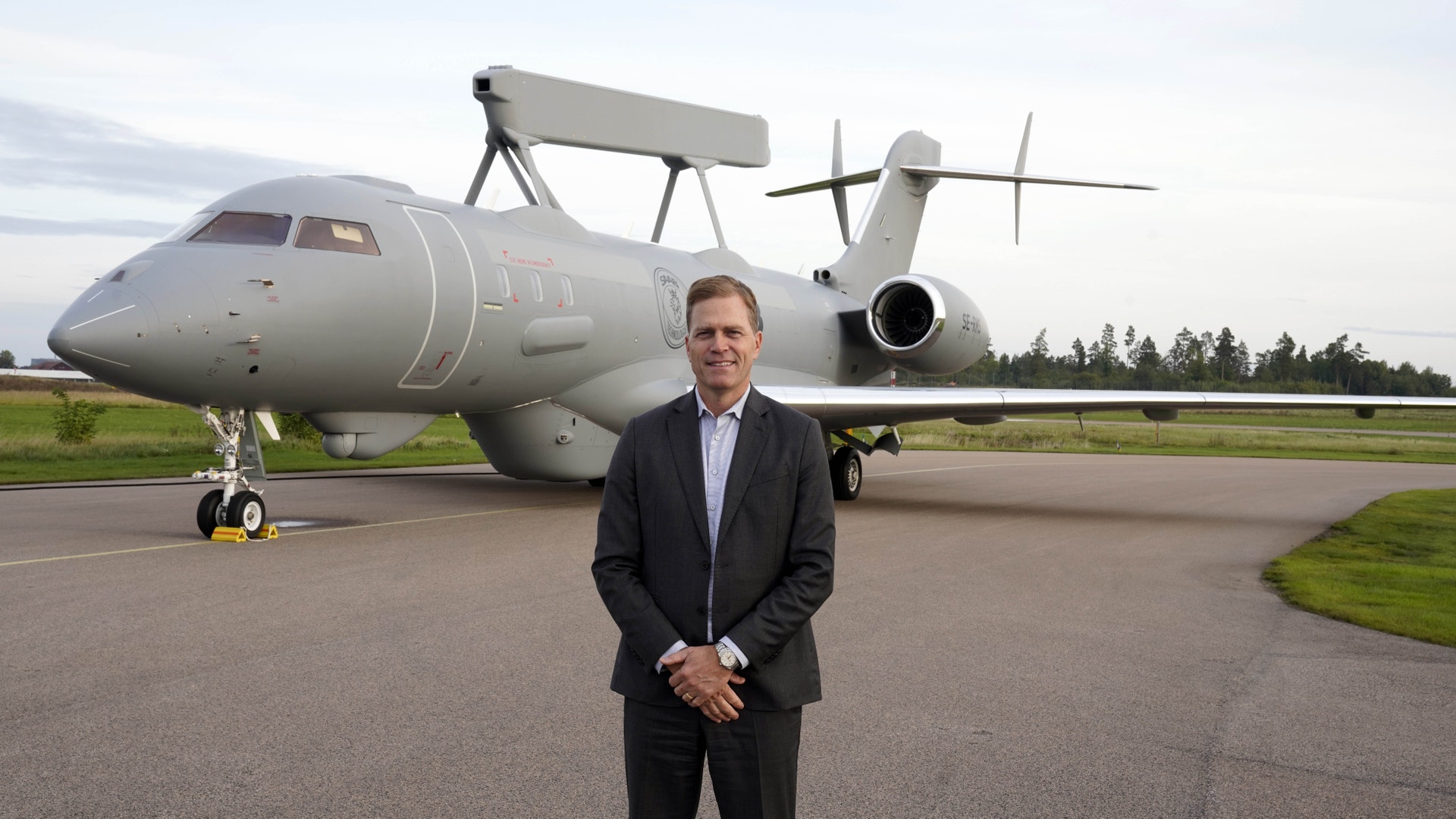 En man i mörk kavaj står framför ett grått flygplan med radarantenn på ryggen. Planet står på en flygplats.