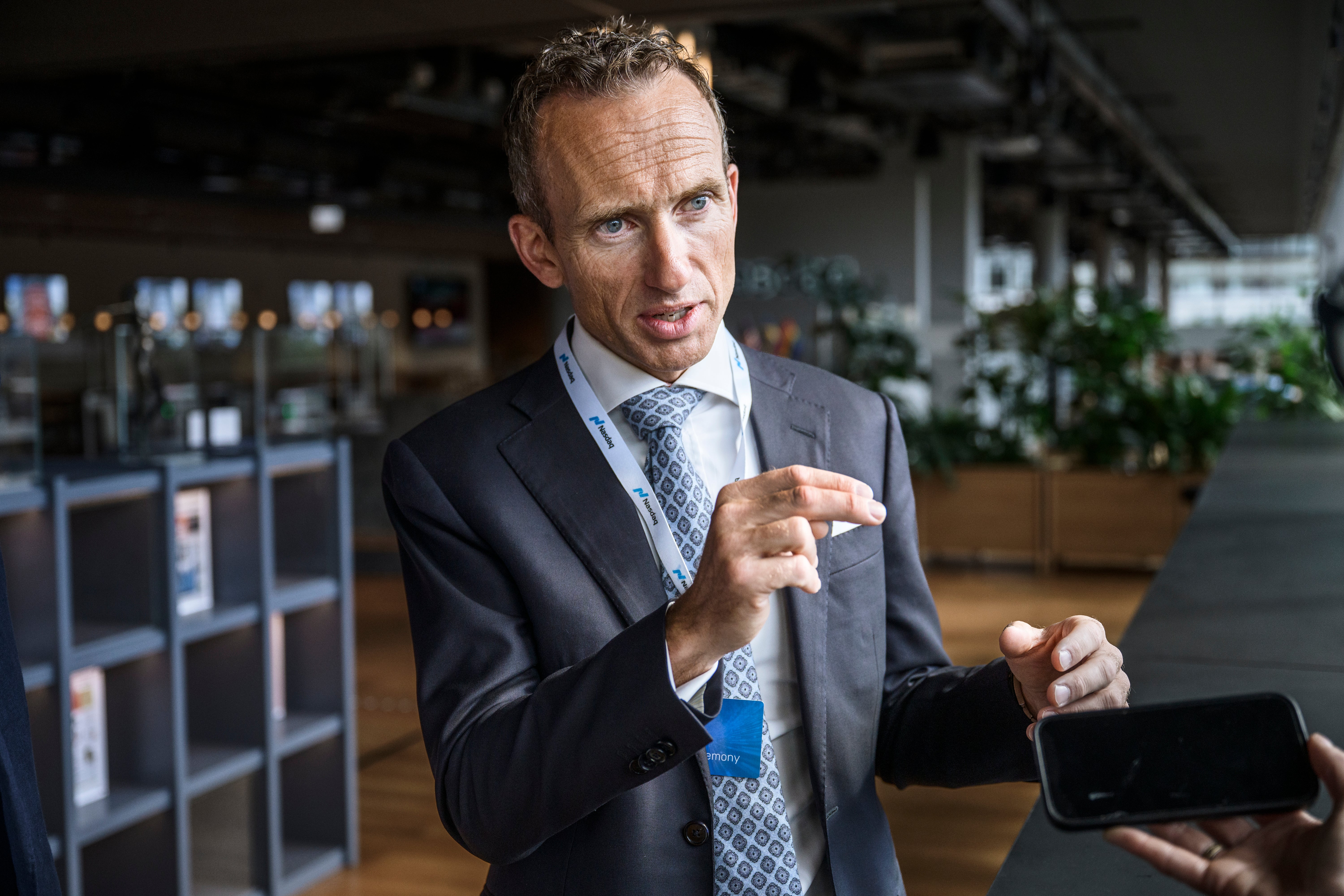 Man i kostym och slips håller en smartphone och gestikulerar medan han presenterar något i en modern kontorsmiljö.