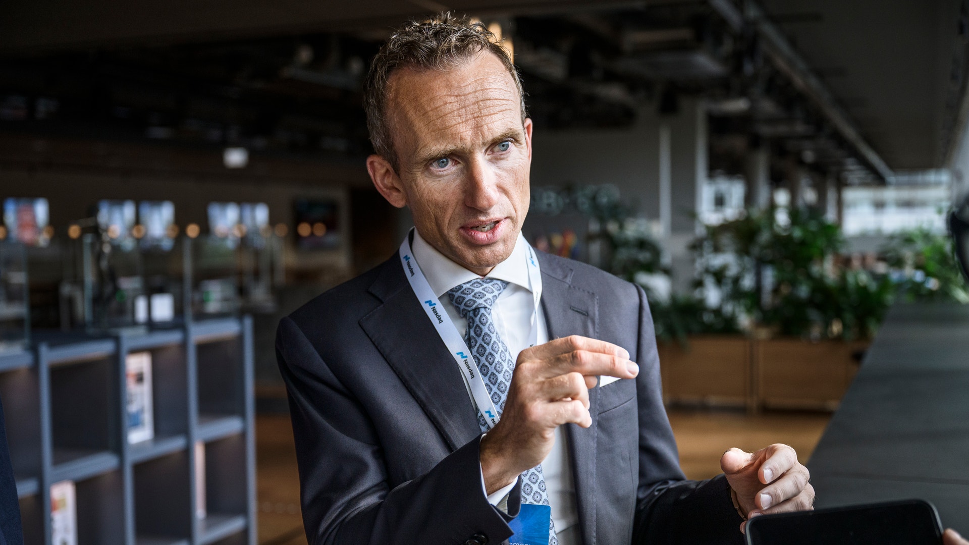 Man i kostym och slips håller en smartphone och gestikulerar medan han presenterar något i en modern kontorsmiljö.