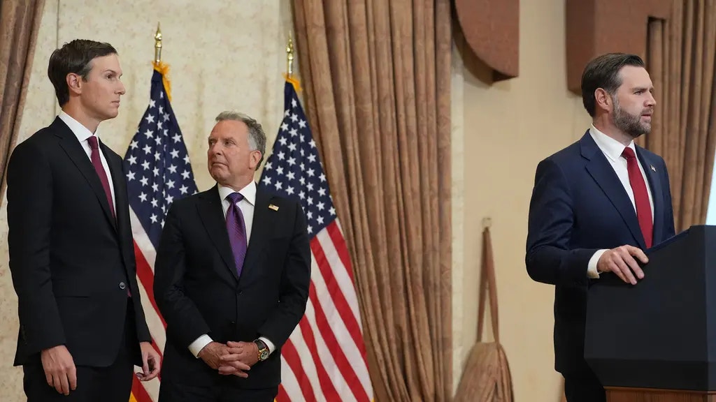 Tre män i formell affärsdräkt står i ett officiellt regeringsrum med amerikanska flaggor i bakgrunden.