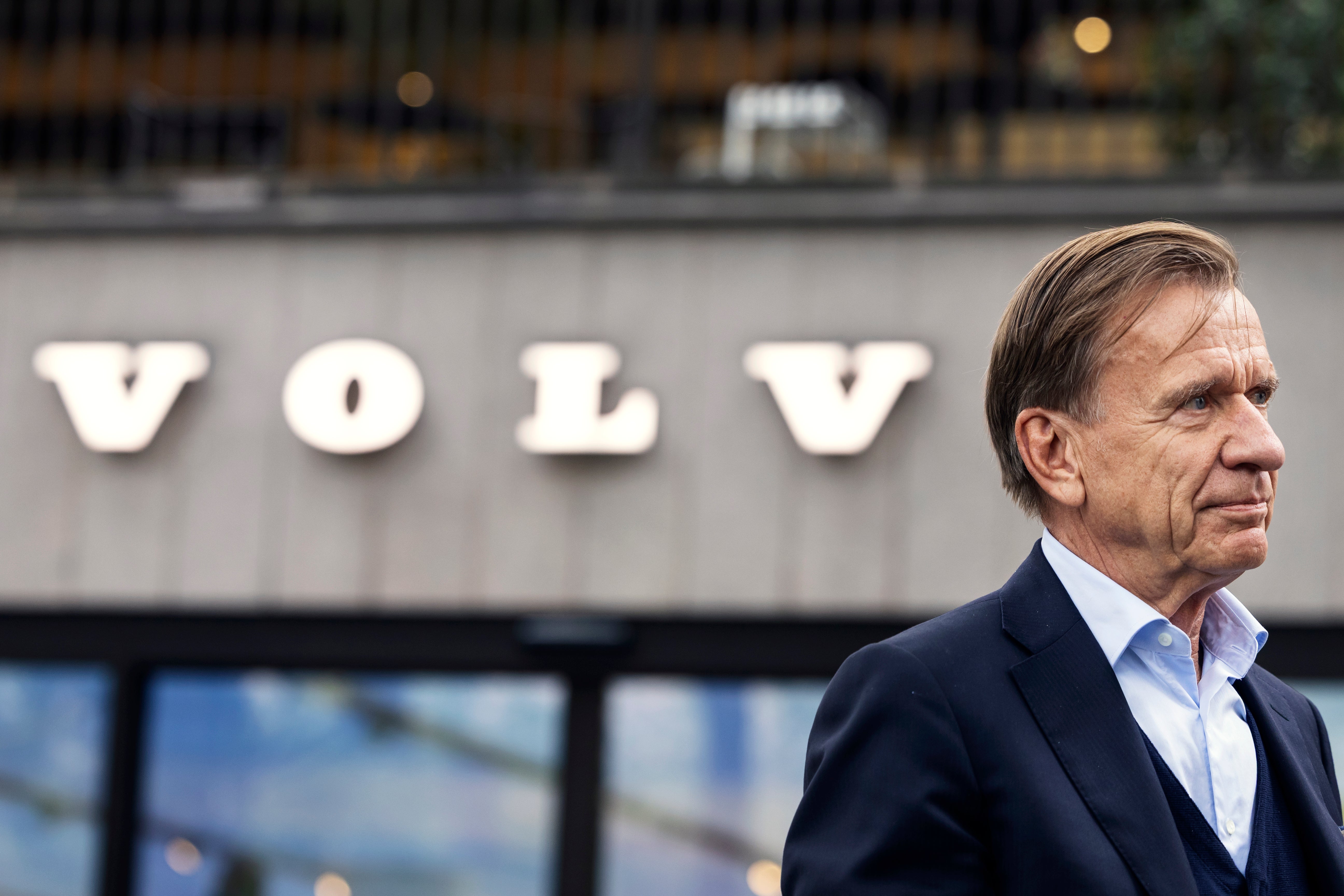 En man i kostym står utanför en byggnad med Volvos logotyp i bakgrunden.