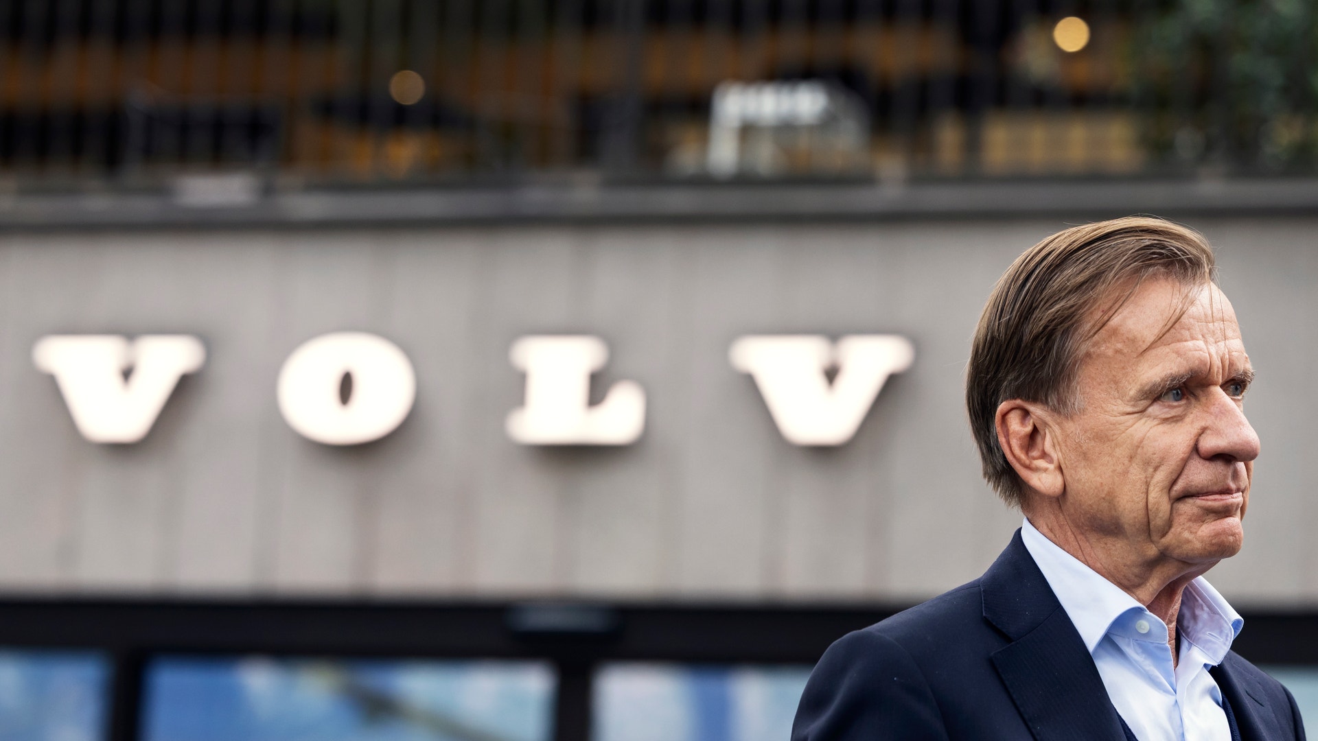 En man i kostym står utanför en byggnad med Volvos logotyp i bakgrunden.