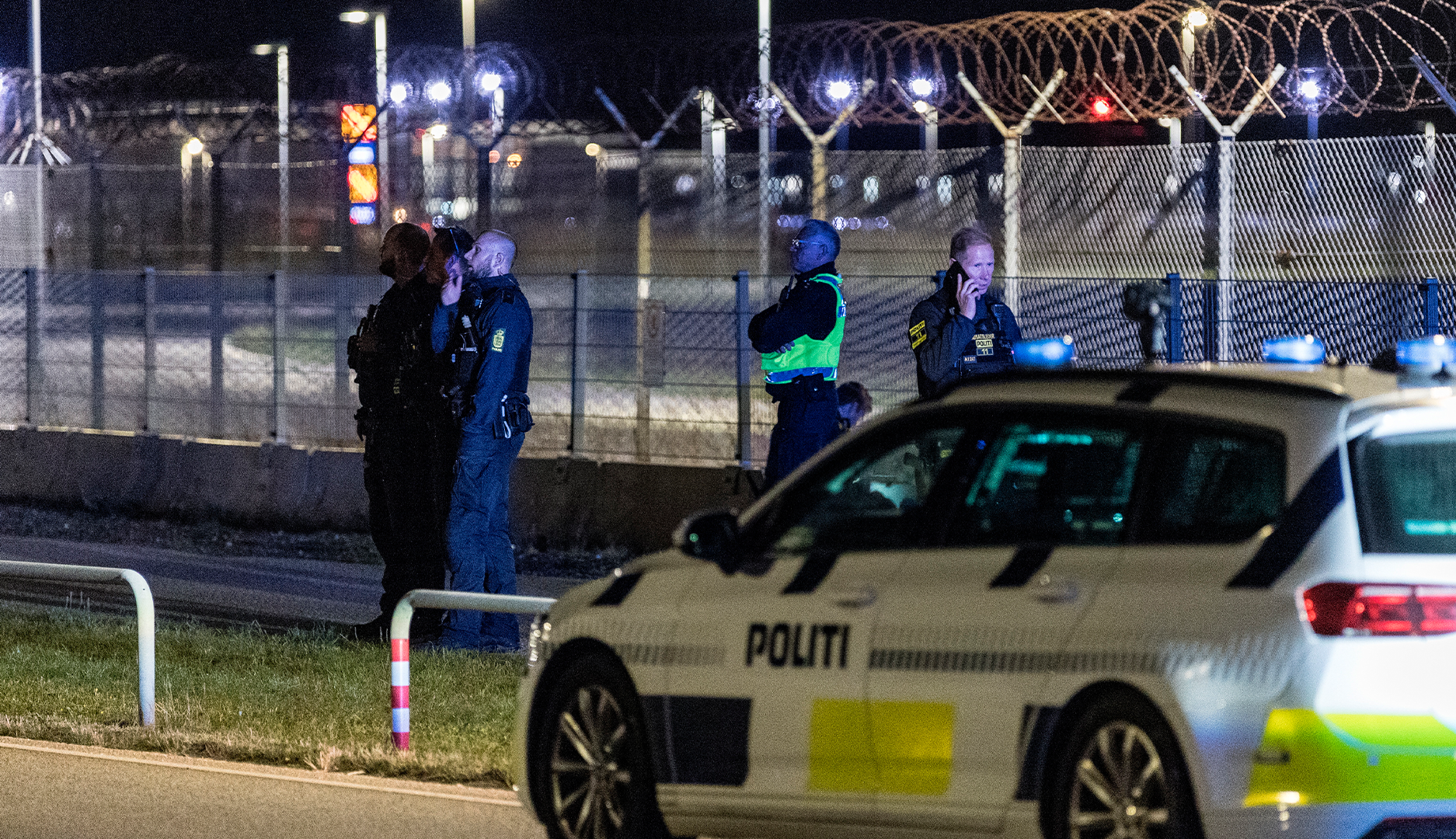 En dansk polisbil och poliser vid ett säkrat område med högt staket och taggtråd, fotograferat på natten.