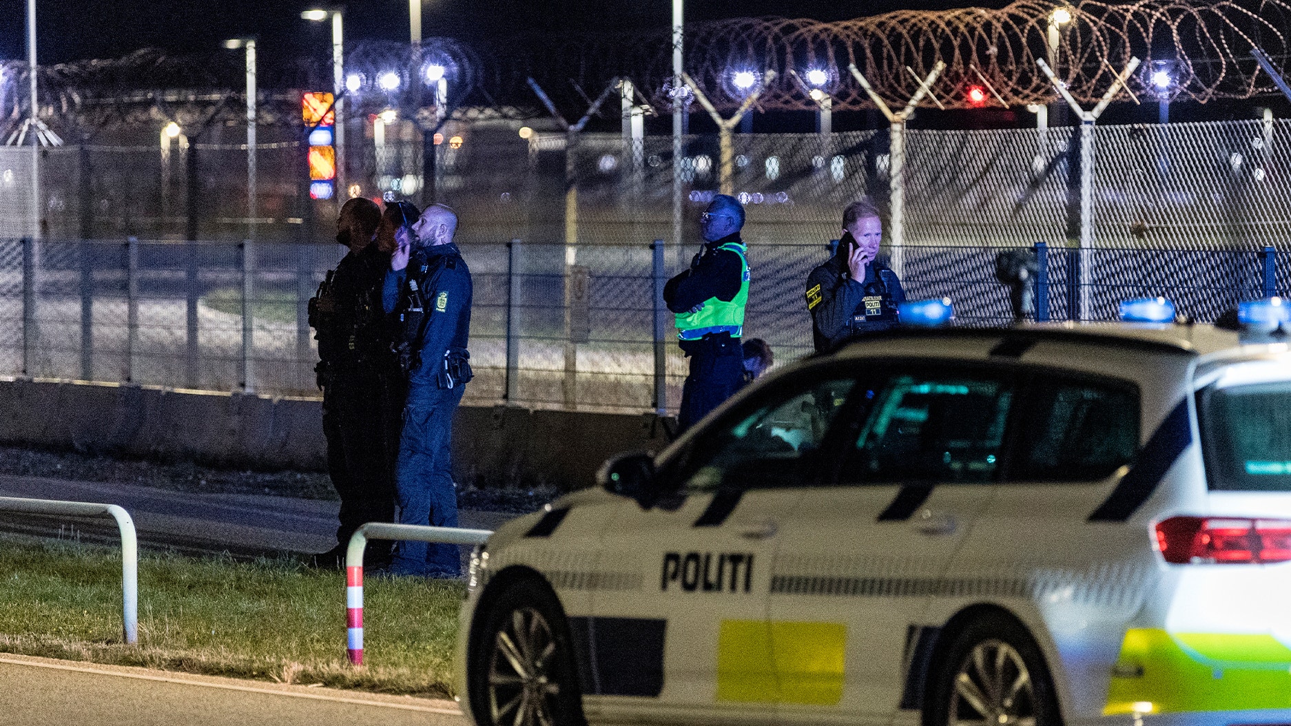 En dansk polisbil och poliser vid ett säkrat område med högt staket och taggtråd, fotograferat på natten.