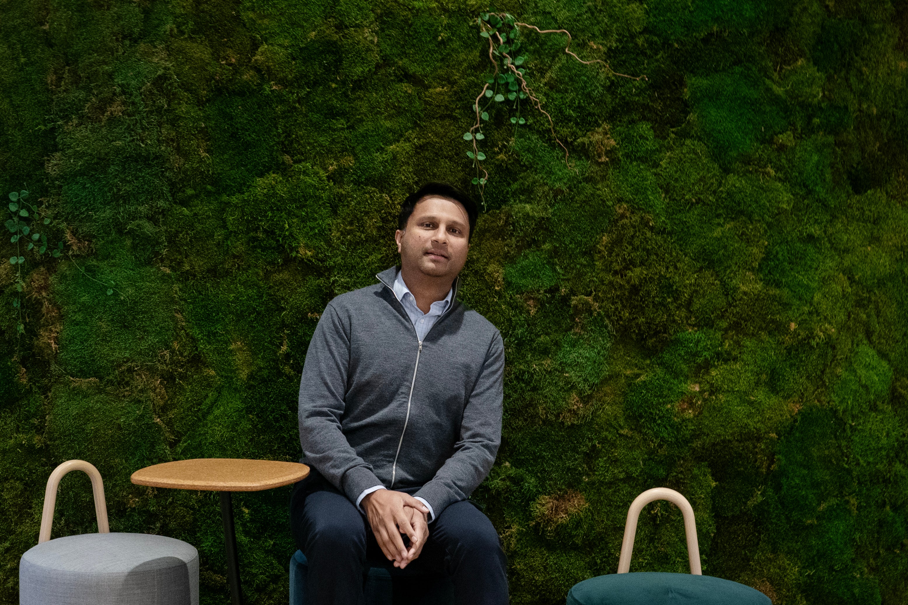 En man i mörkgrå hoodie sitter på en modern stol framför en vägg täckt med levande mossa. Han har en lugn och självsäker blick och håller händerna korsade över sitt knä. Bakgrunden visar en rik grön mossvägg med naturliga variationer i färg och textur.