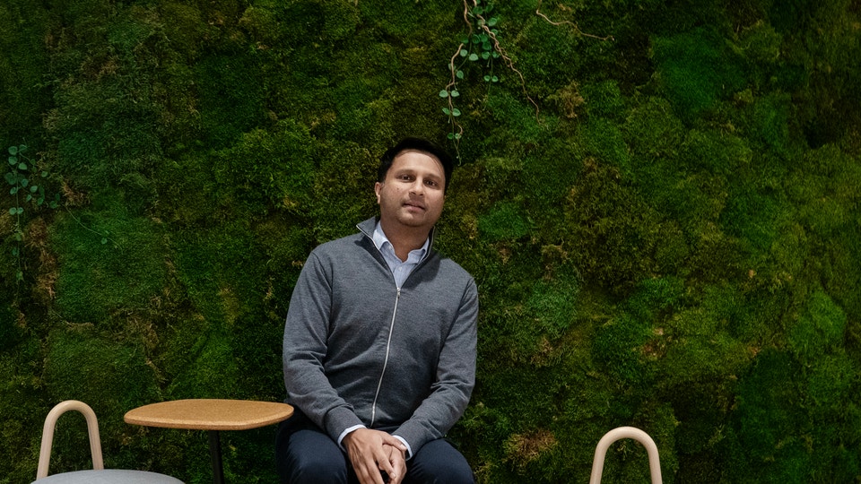 En man i mörkgrå hoodie sitter på en modern stol framför en vägg täckt med levande mossa. Han har en lugn och självsäker blick och håller händerna korsade över sitt knä. Bakgrunden visar en rik grön mossvägg med naturliga variationer i färg och textur.