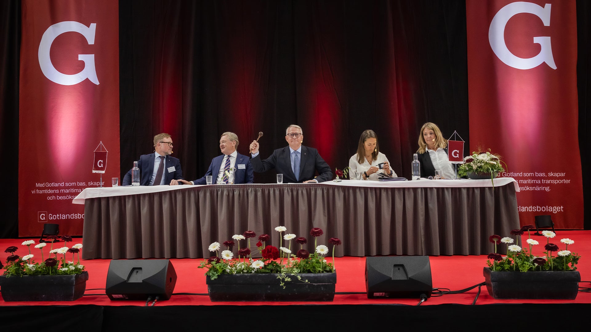 Fem personer sitter vid ett bord på en scen under en paneldiskussion. Bakgrunden är röd med stora G-logotyper, och framför scenen finns blomsterarrangemang.