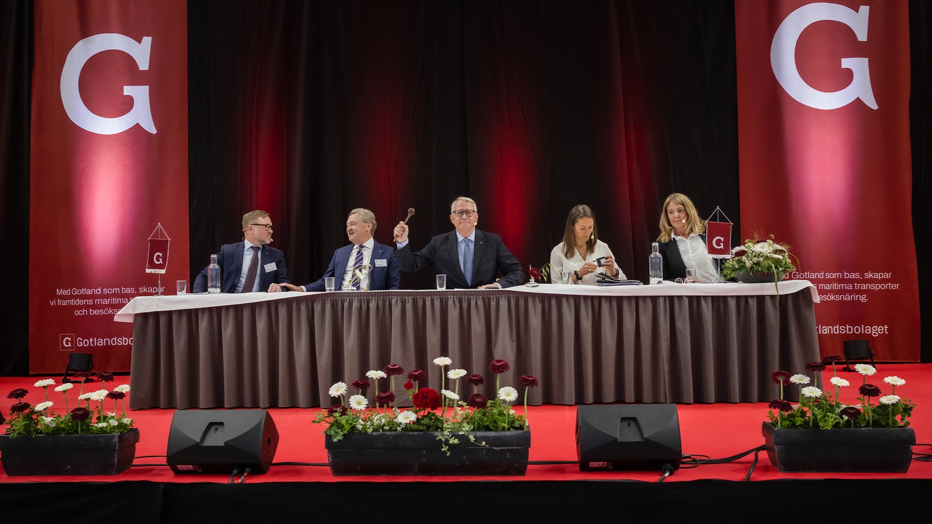 Fem personer sitter vid ett bord på en scen under en paneldiskussion. Bakgrunden är röd med stora G-logotyper, och framför scenen finns blomsterarrangemang.