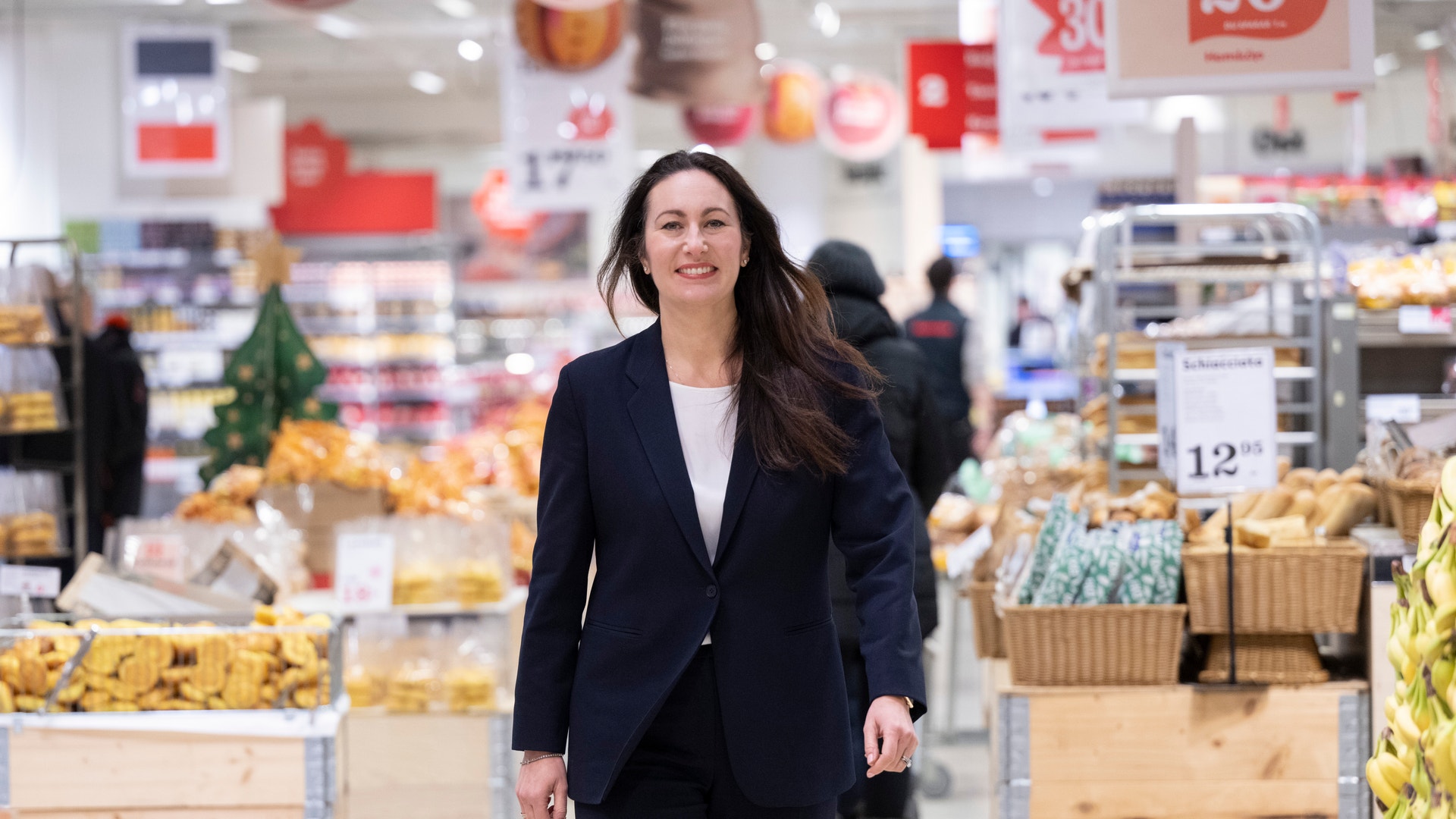 Kvinna i mörk kostym går genom en matbutik mellan hyllor med varor.