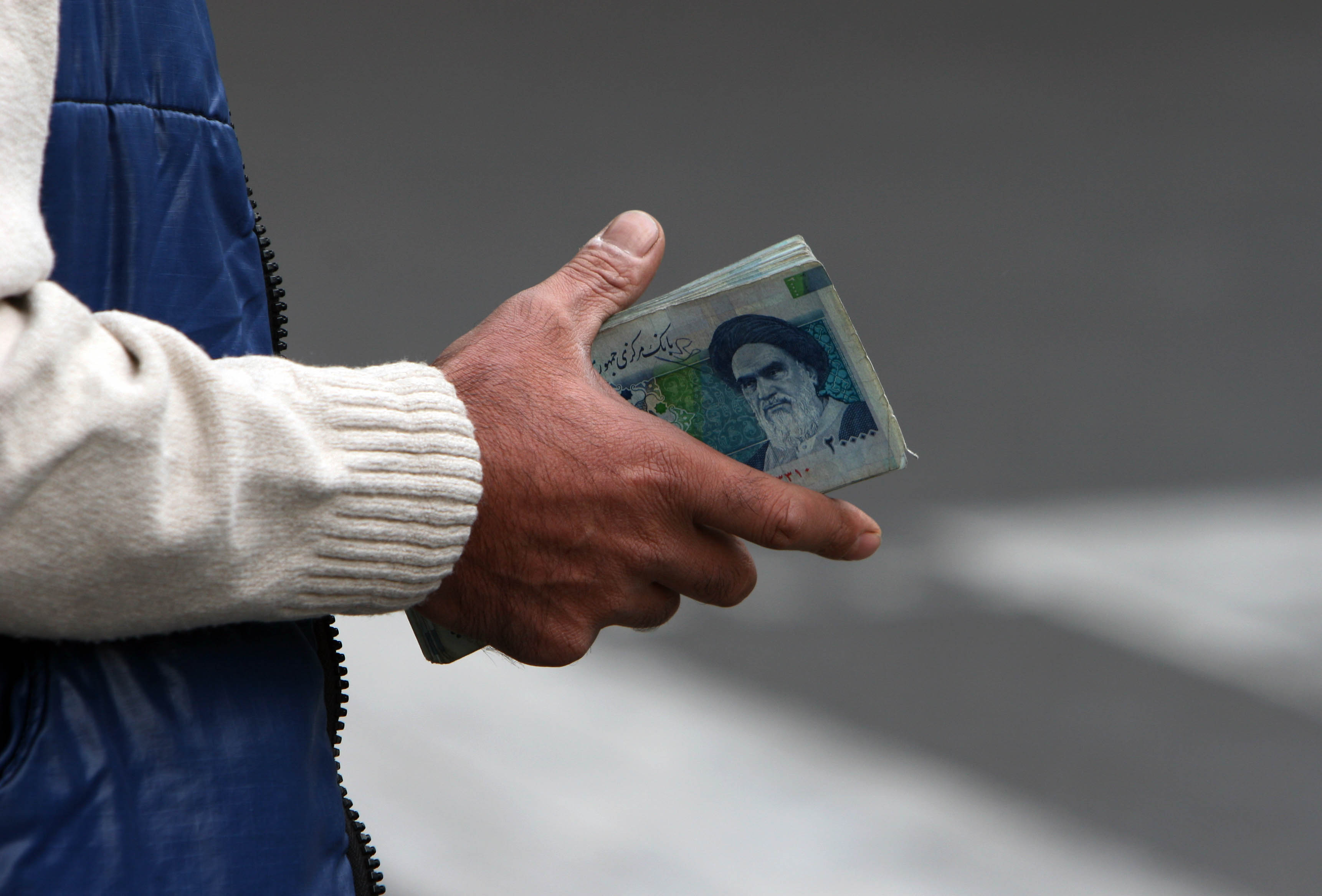 En hand håller i en sedel. Personen bär en vit stickad tröja och en blå jacka.Iran, valuta, Rial.