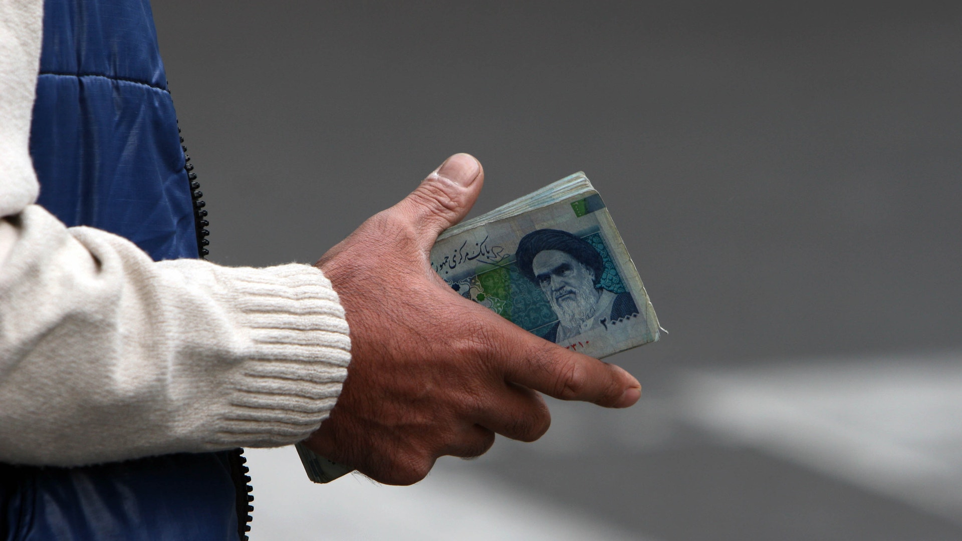En hand håller i en sedel. Personen bär en vit stickad tröja och en blå jacka.Iran, valuta, Rial.