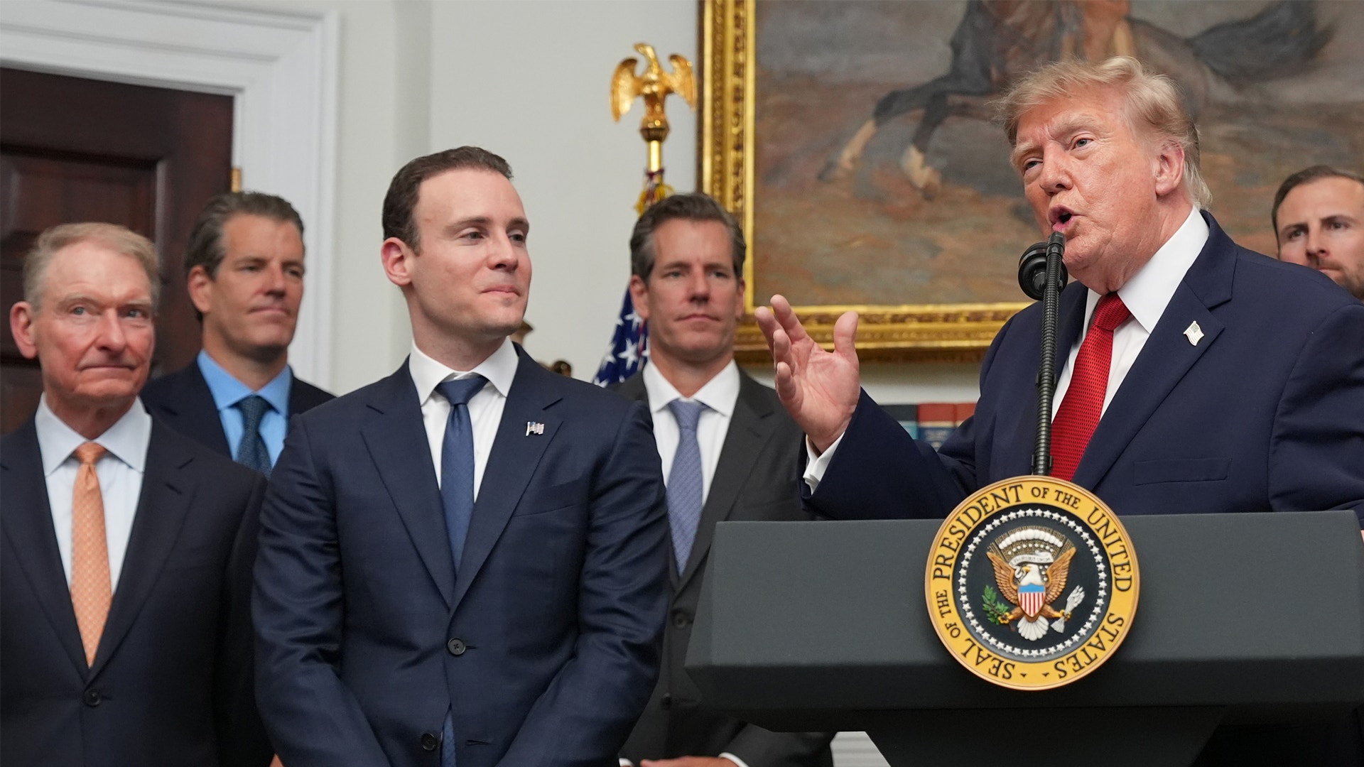 En man i röd slips talar vid ett podium med presidentens sigill i Vita huset, omgiven av flera män i formella kostymer.
