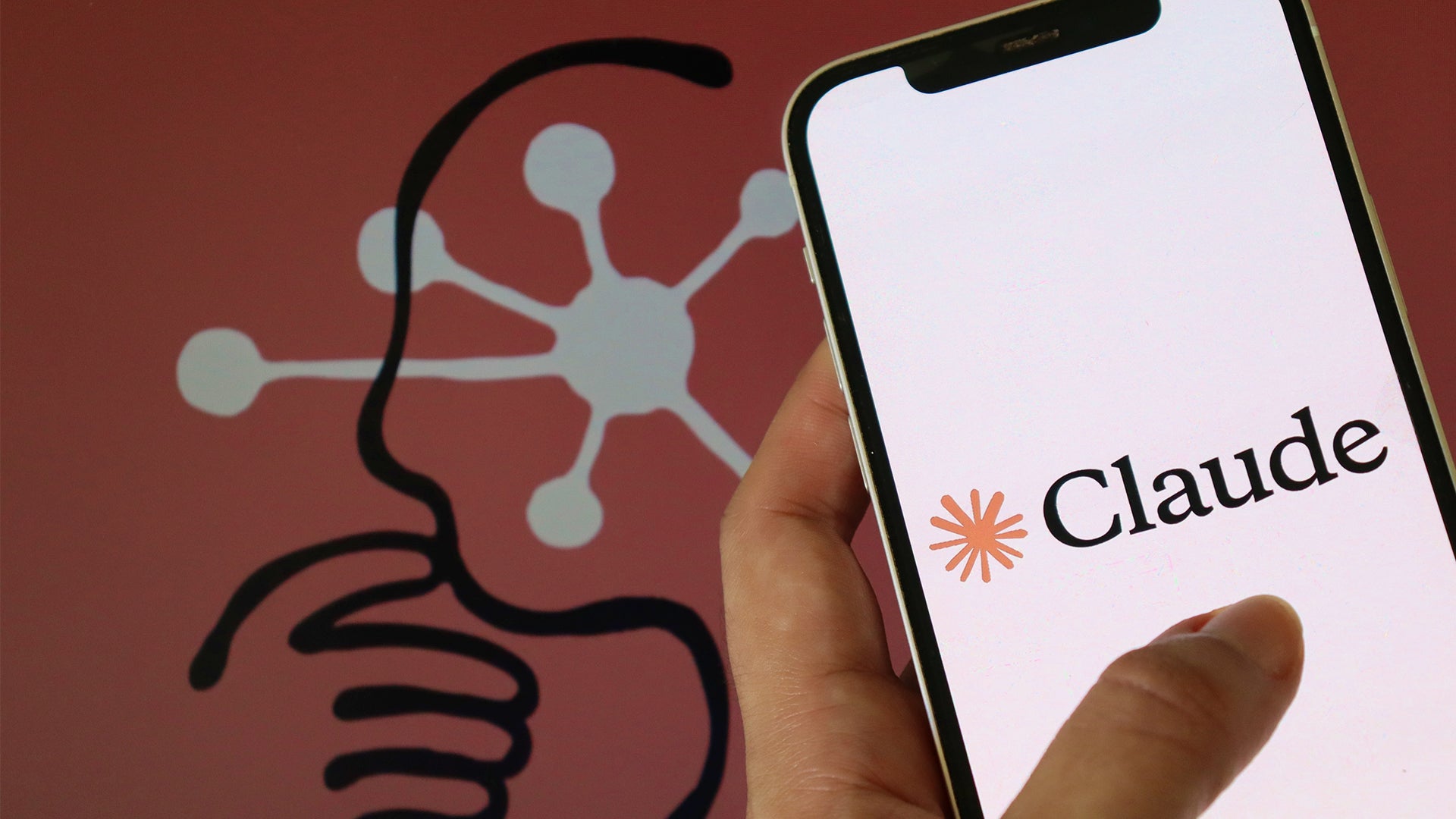 En hand håller en smartphone med Claude-logon på skärmen. I bakgrunden syns en illustration av en hjärna med nätverksstrukturer som representerar artificiell intelligens.