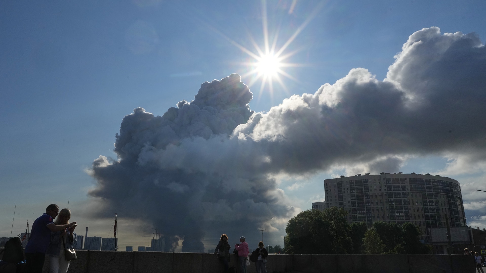Människor står på en takterrass och tittar upp mot en dramatisk himmel med stora moln och starkt solljus. Moderna bostadshus syns i bakgrunden.