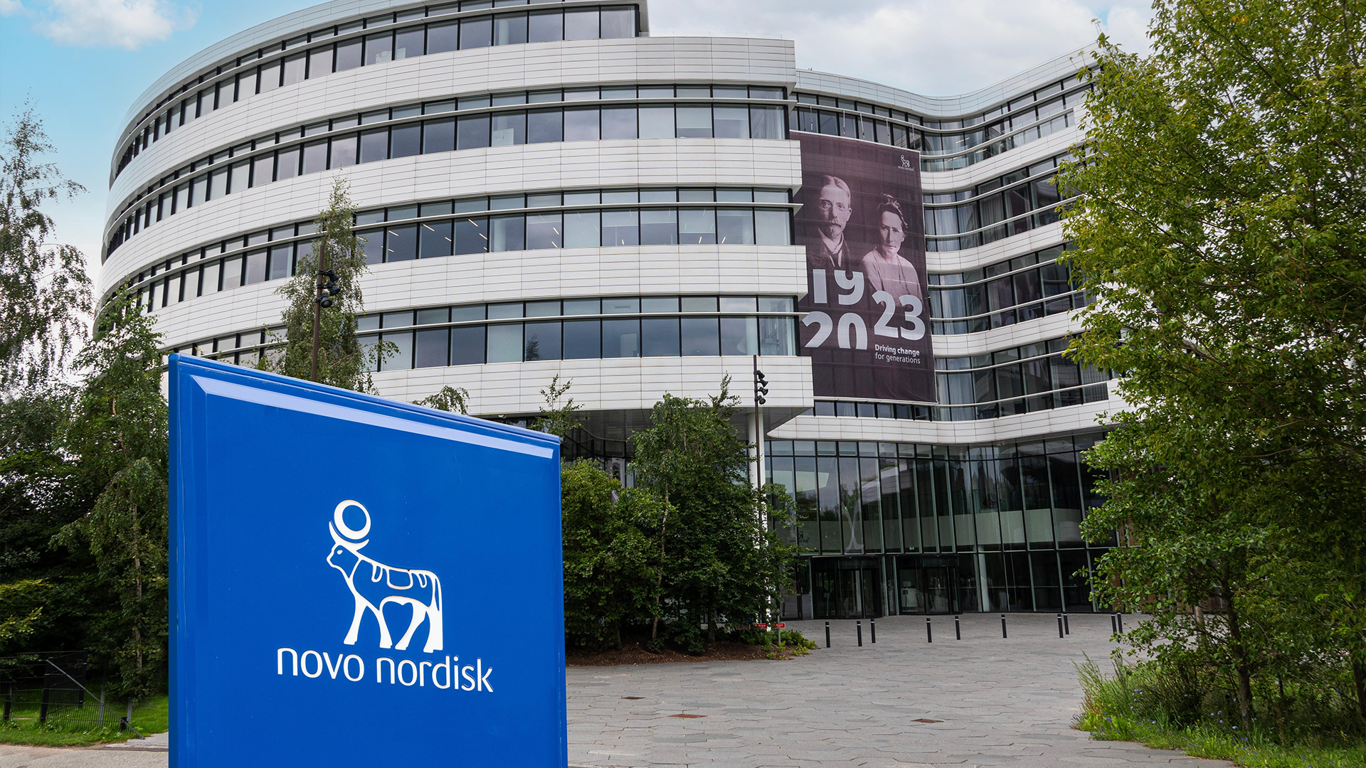 Novo Nordisks huvudkontor, en modern rundad byggnad med vita och grå fasader, med blå skylt med företagslogon i förgrunden.