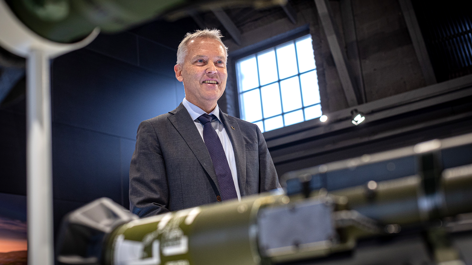 En man i kostym och slips står i en industriell miljö med stora fönster i bakgrunden.