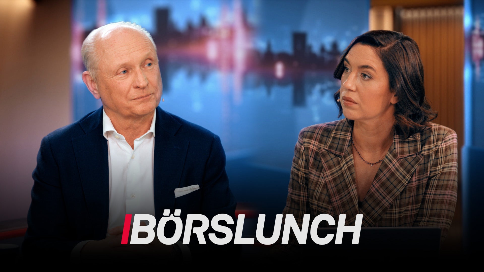 En man och en kvinna sitter vid ett skrivbord framför en stadssilhuett. Bilden är från programmet Börslunch.