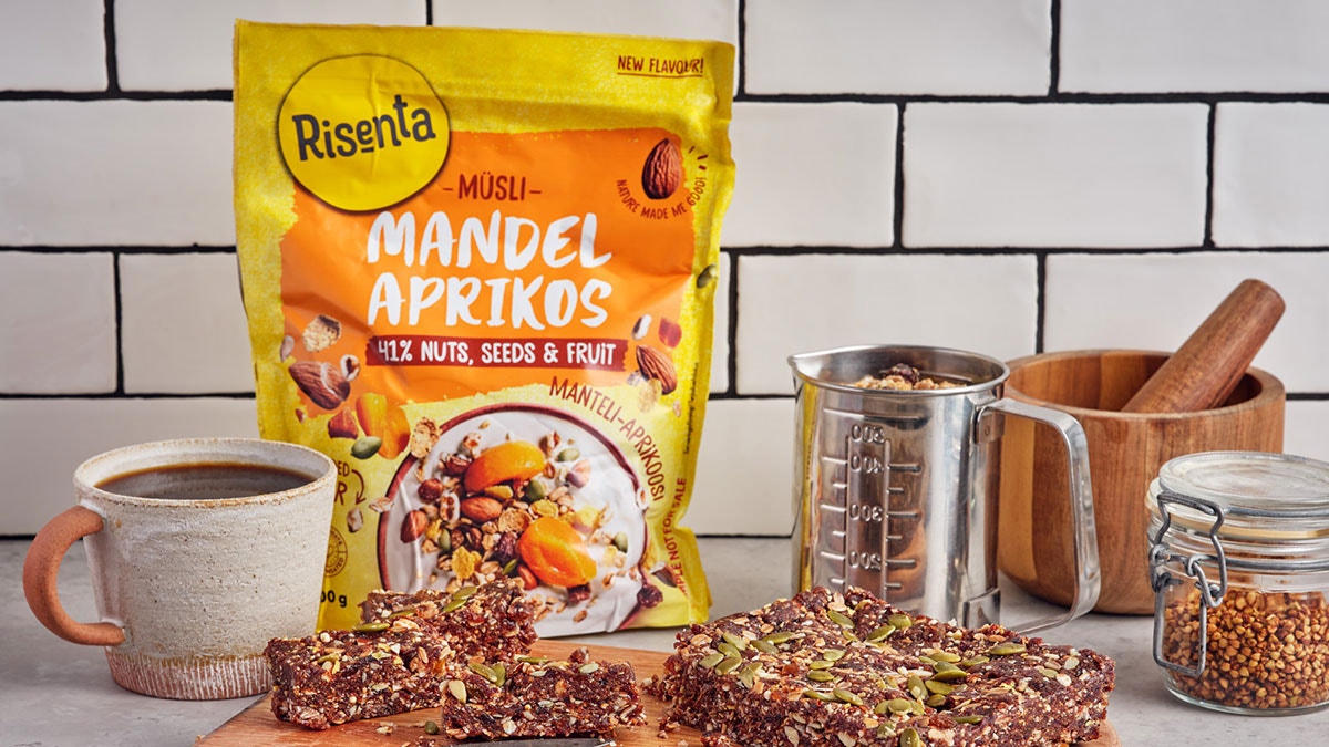 Risenta müsli-paket med mandel och aprikoser omgivet av keramikmugg, glasburkar med spannmål och träkärl på vit kakelvägg.