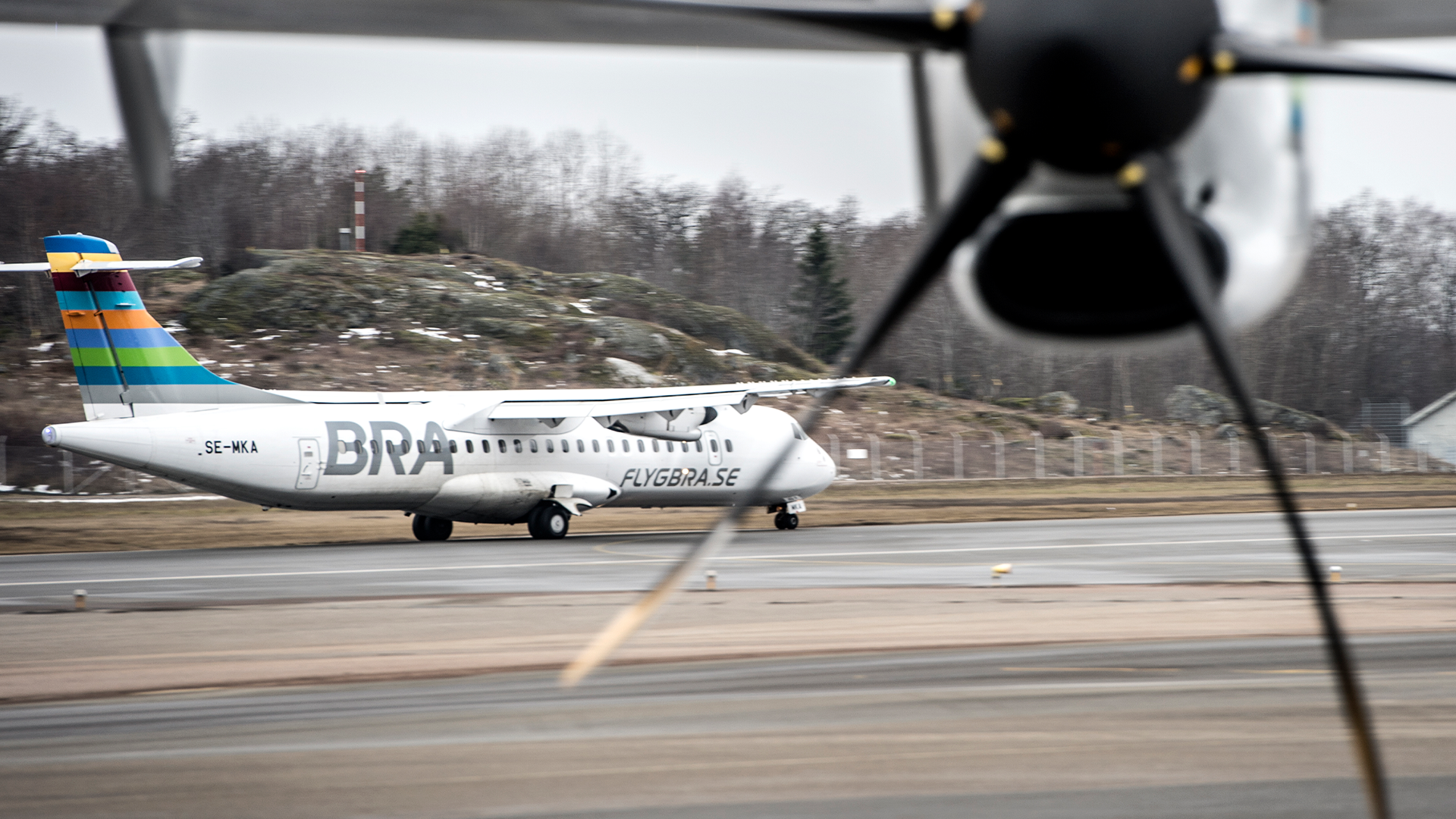 Ett passagerarflygplan på landningsbanan, fotograferat från ett annat flygplan. Flygplanet har blå och orange stripes på stjärten och rullar på asfalten med skogar och kullar i bakgrunden.