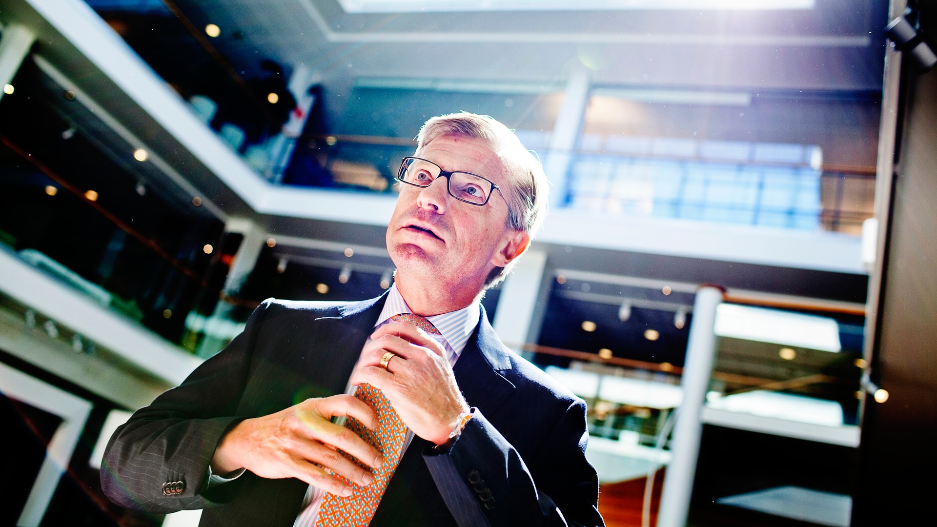 En man i affärskläder och glasögon står i en modern kontorsmiljö och gestikulerar medan han talar.