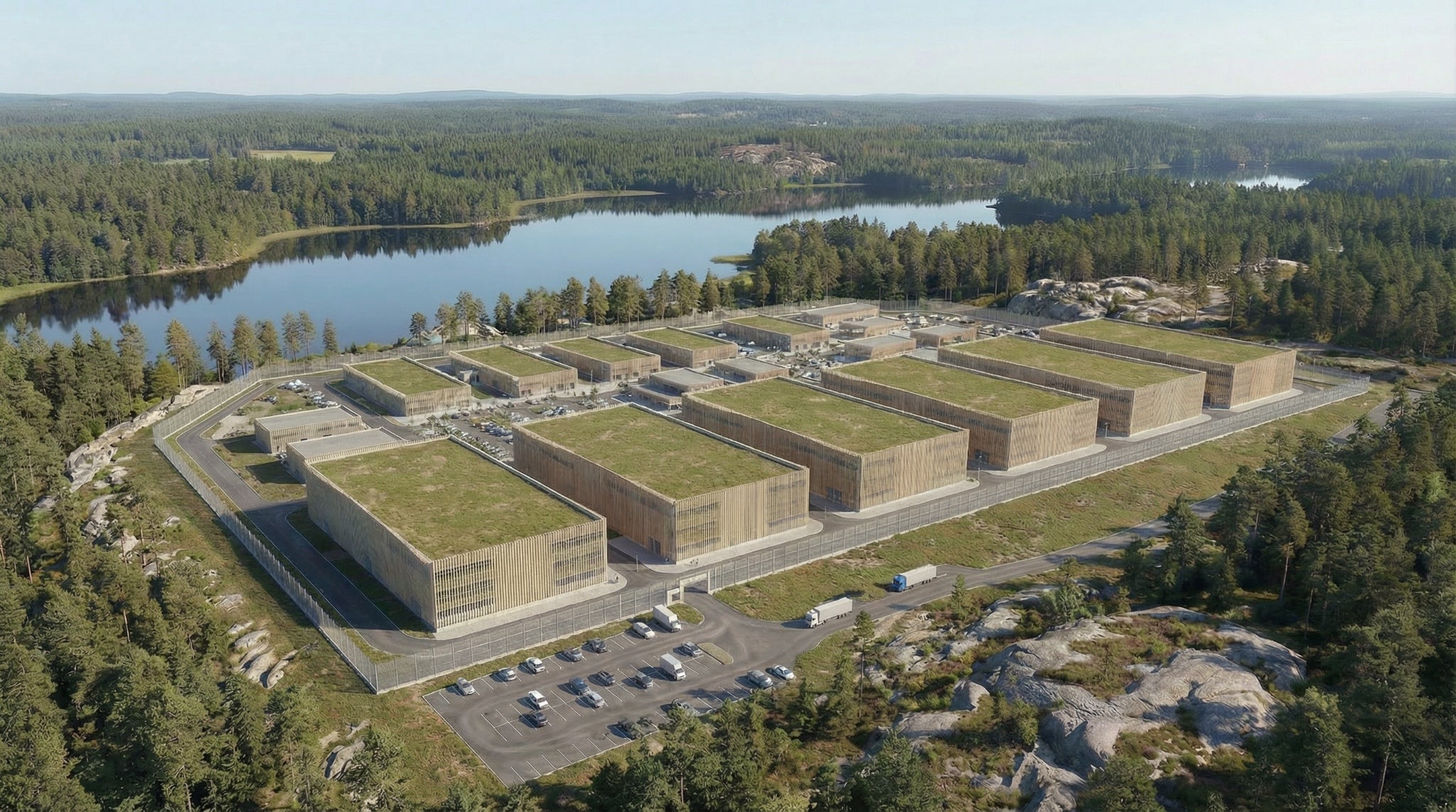 Flygfoto över en modern byggnad med gröna tak omgiven av skog och vatten.