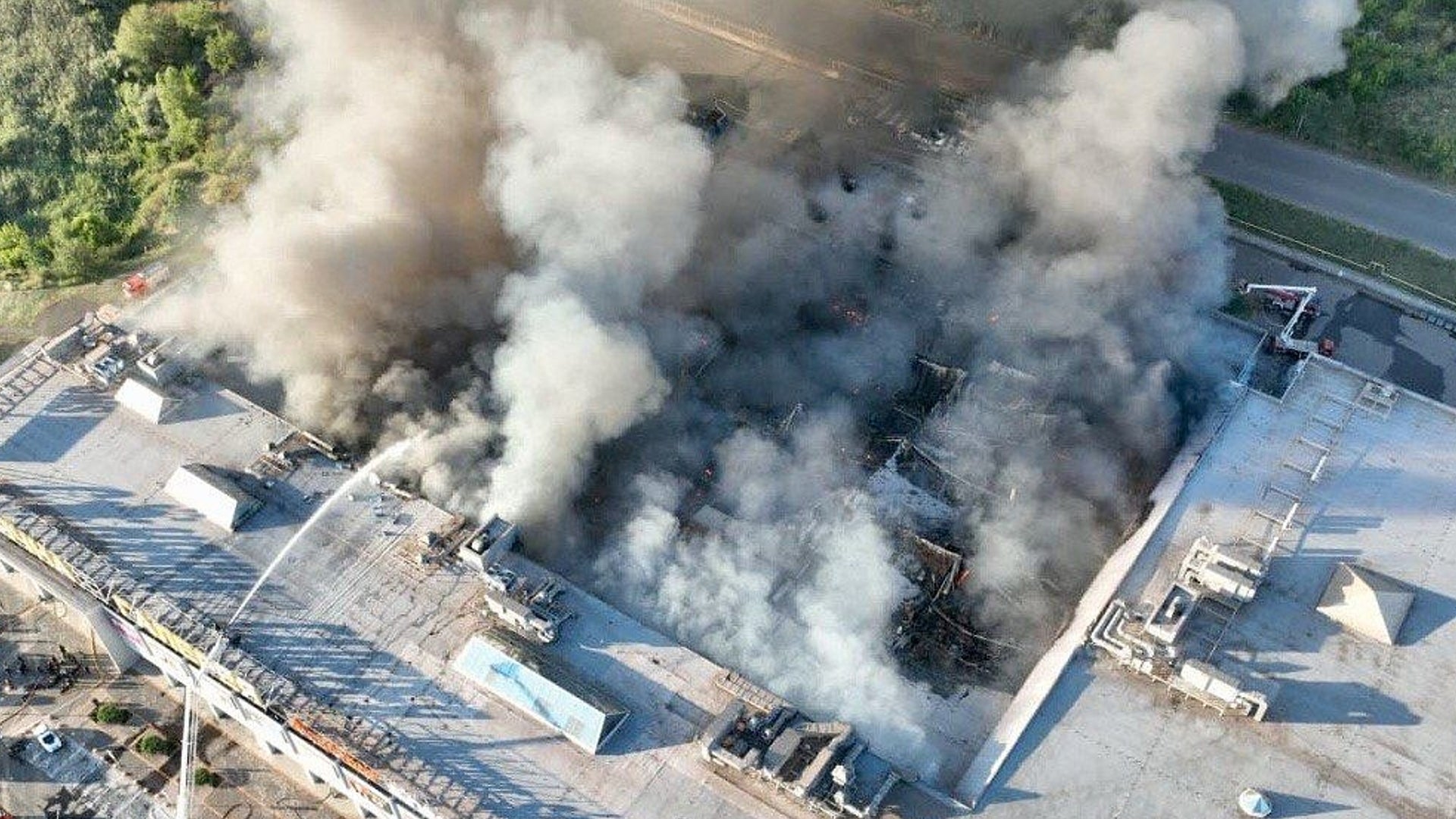 Flygfoto över en industribyggnad som är i brand med tjock vit och svart rök som stiger från taket.