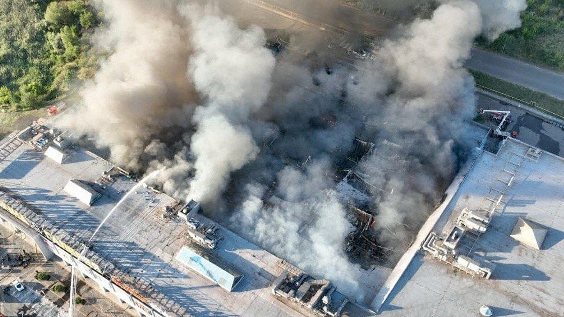 Flygfoto över en industribyggnad som är i brand med tjock vit och svart rök som stiger från taket.