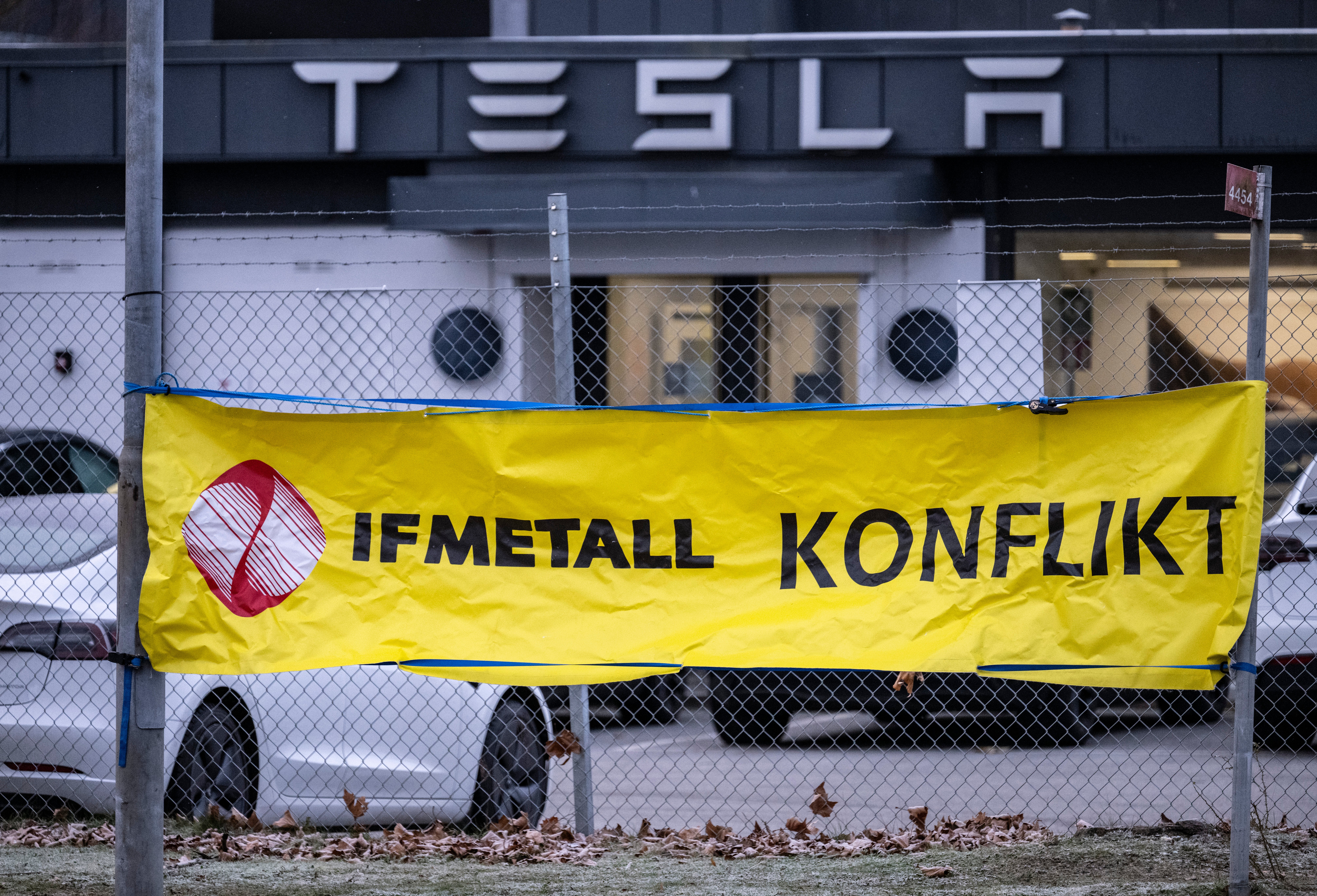 Gul banner med texten IFMETALL KONFLIKT hängande på ett staket framför en Tesla-byggnad.