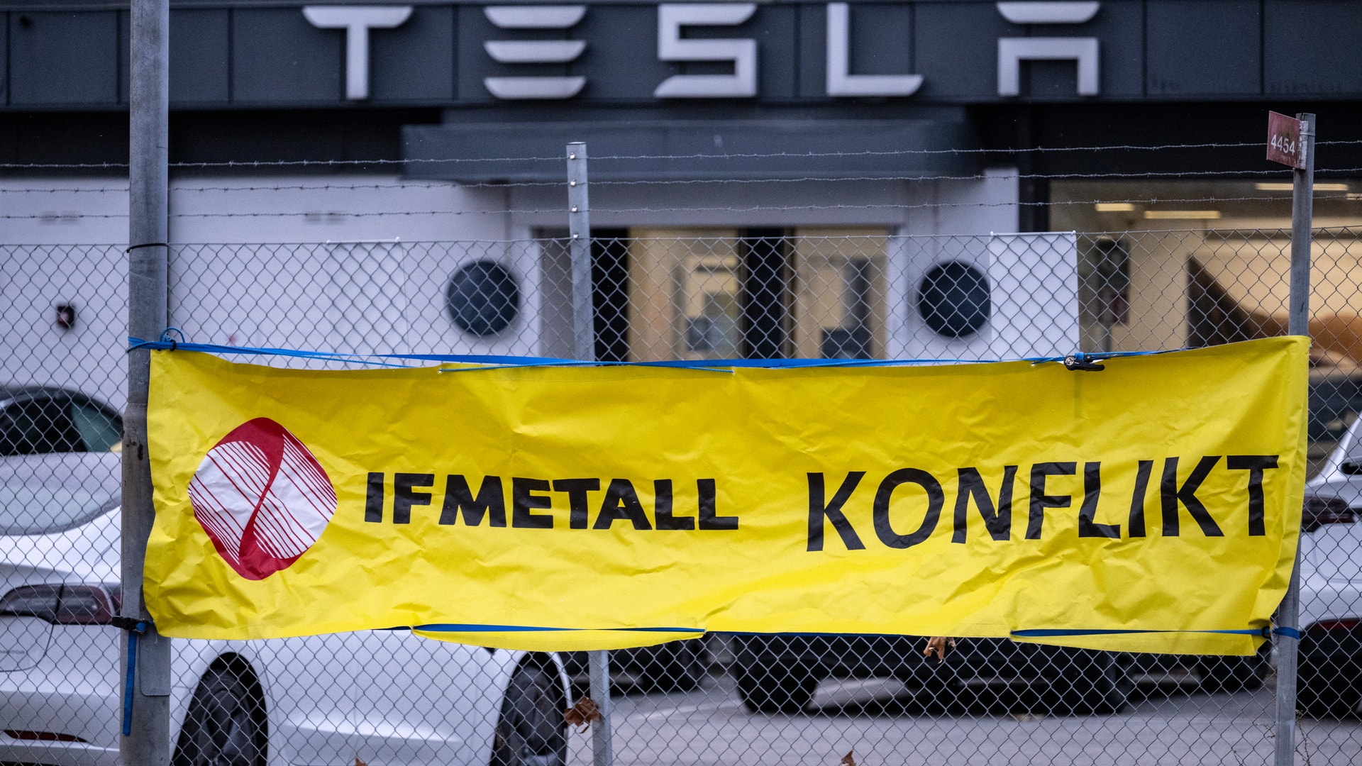 Gul banner med texten IFMETALL KONFLIKT hängande på ett staket framför en Tesla-byggnad.