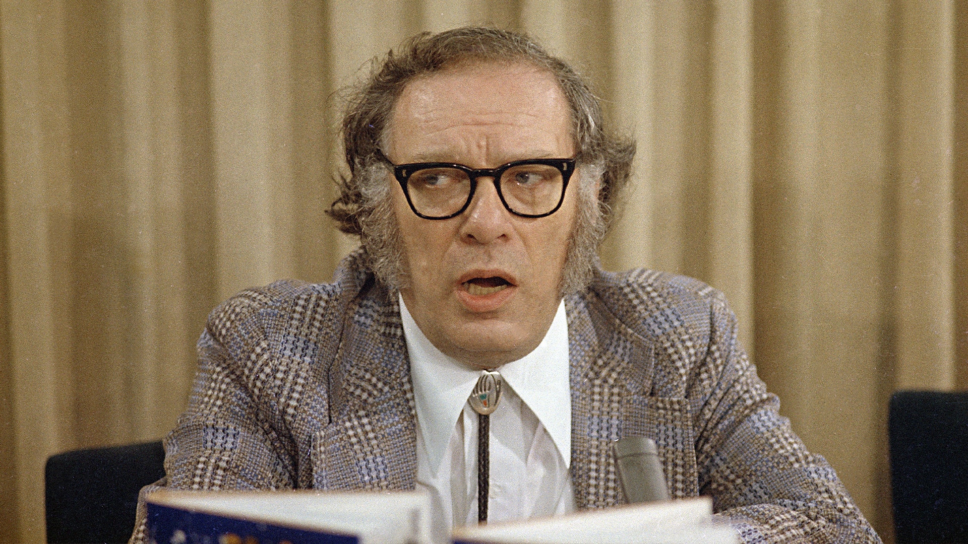 En man med glasögon och rutigt jackett talar vid en mikrofon under en formell tillställning.