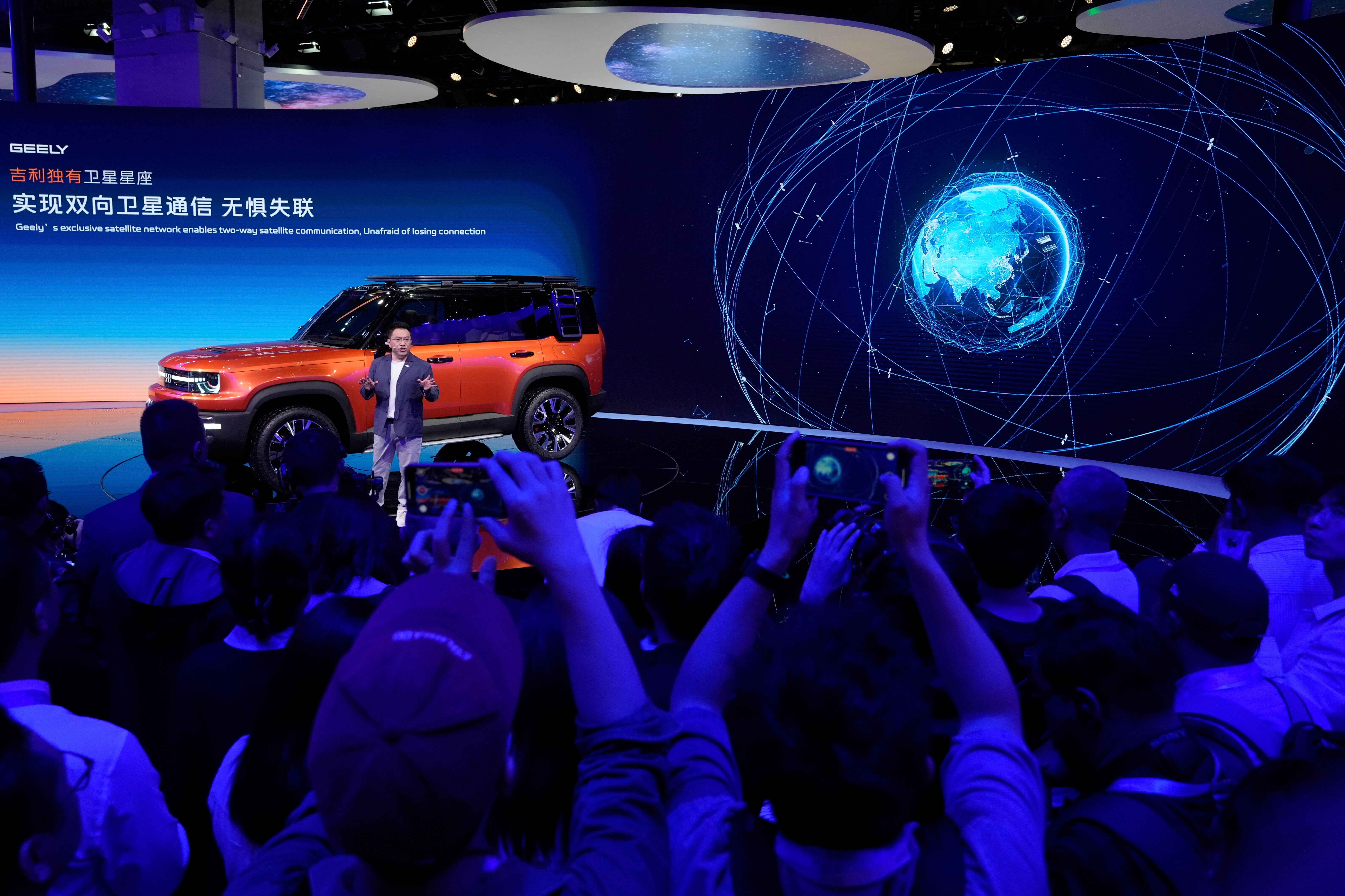 Presentatör visar upp en orange SUV på en belyst scen medan publik fotograferar med sina telefoner.