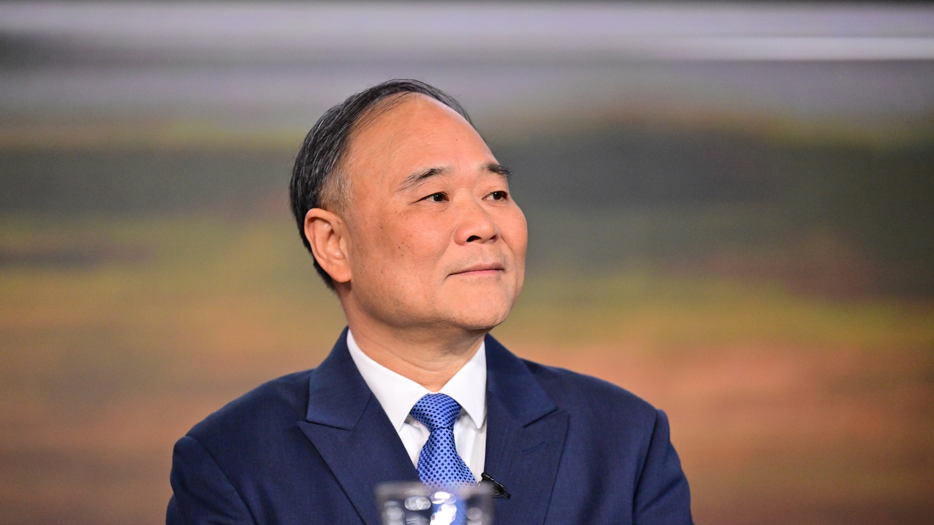 Porträtt av en man i mörkblå kostym och blå slips som sitter vid en mikrofon.