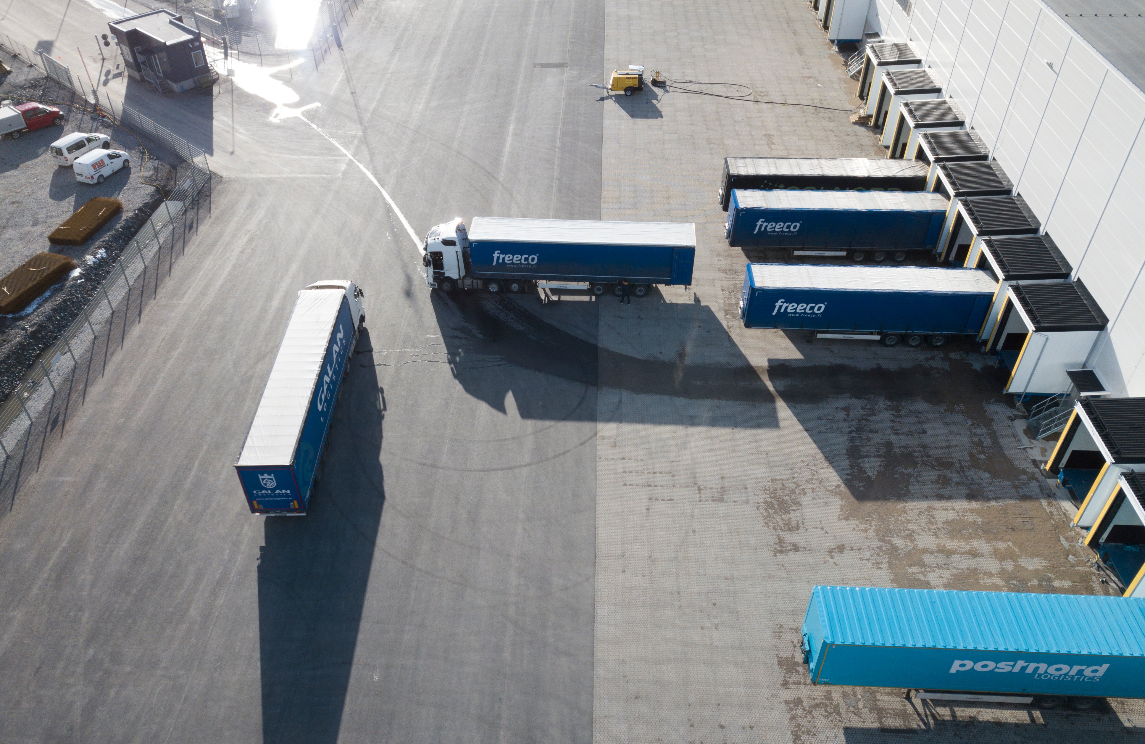 Flygfoto över ett logistikcentrum med flera blå lastbilar parkerade på en betongyta tillsammans med lastningsplattformar och containrar.